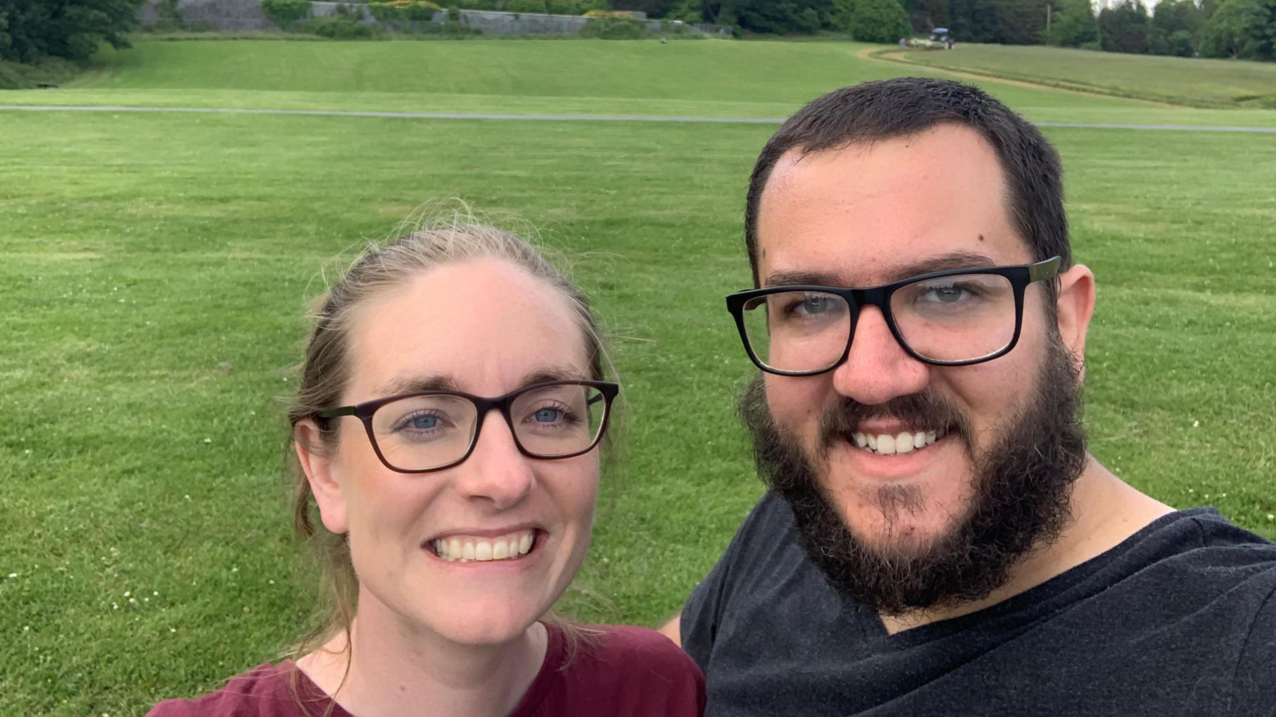 A man and woman (husband and wife) stand on a long lawn in a park smiling into the camera.  The woman is wearing dark rimmed glasses and a red t shirt.  The man beside her has his arm around her.  He is also wearing dark rimmed glasses and he is wearing a dark blue or black t shirt.