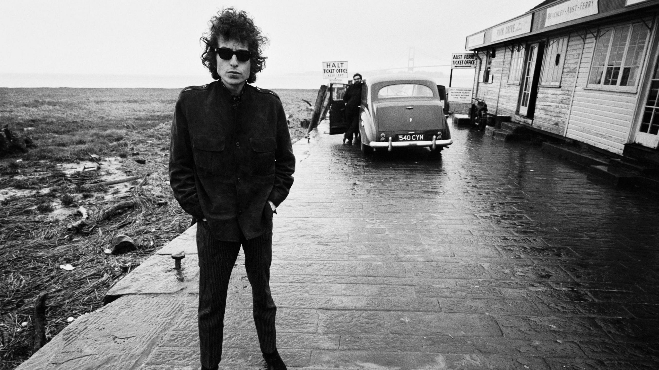 Black and white image of Bob Dylan standing on a platform at the Aust Ferry site. There is a car in the background and a sign that says 'Halt Ticket Office'