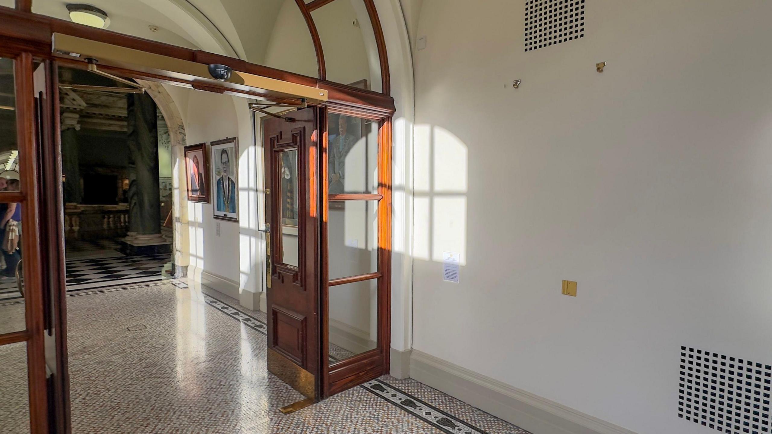 A hallway with wooden double doors, there is a large empty space on the wall where the portrait hung before it was damaged.