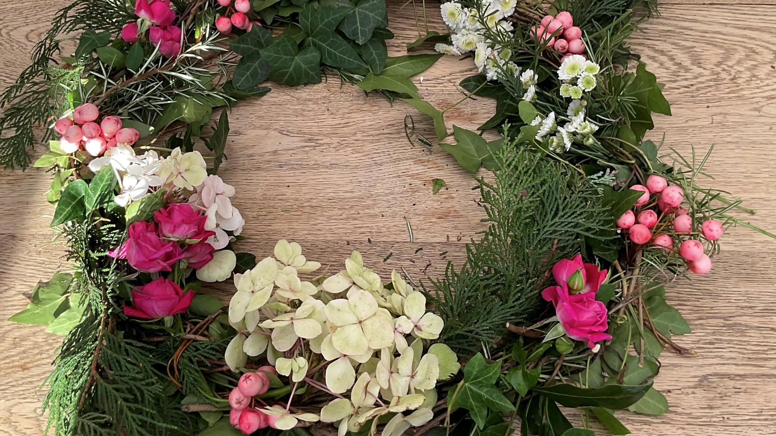 A floral wreathe with white and pink flowers and green foliage sitting on a wooden table top.