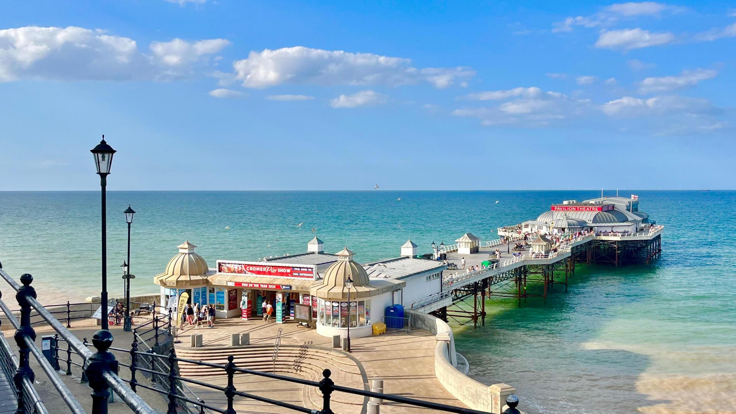 Blue skies with small, fluffy cumulus clouds over the pier at Cromer. Blue and green shades of sea below