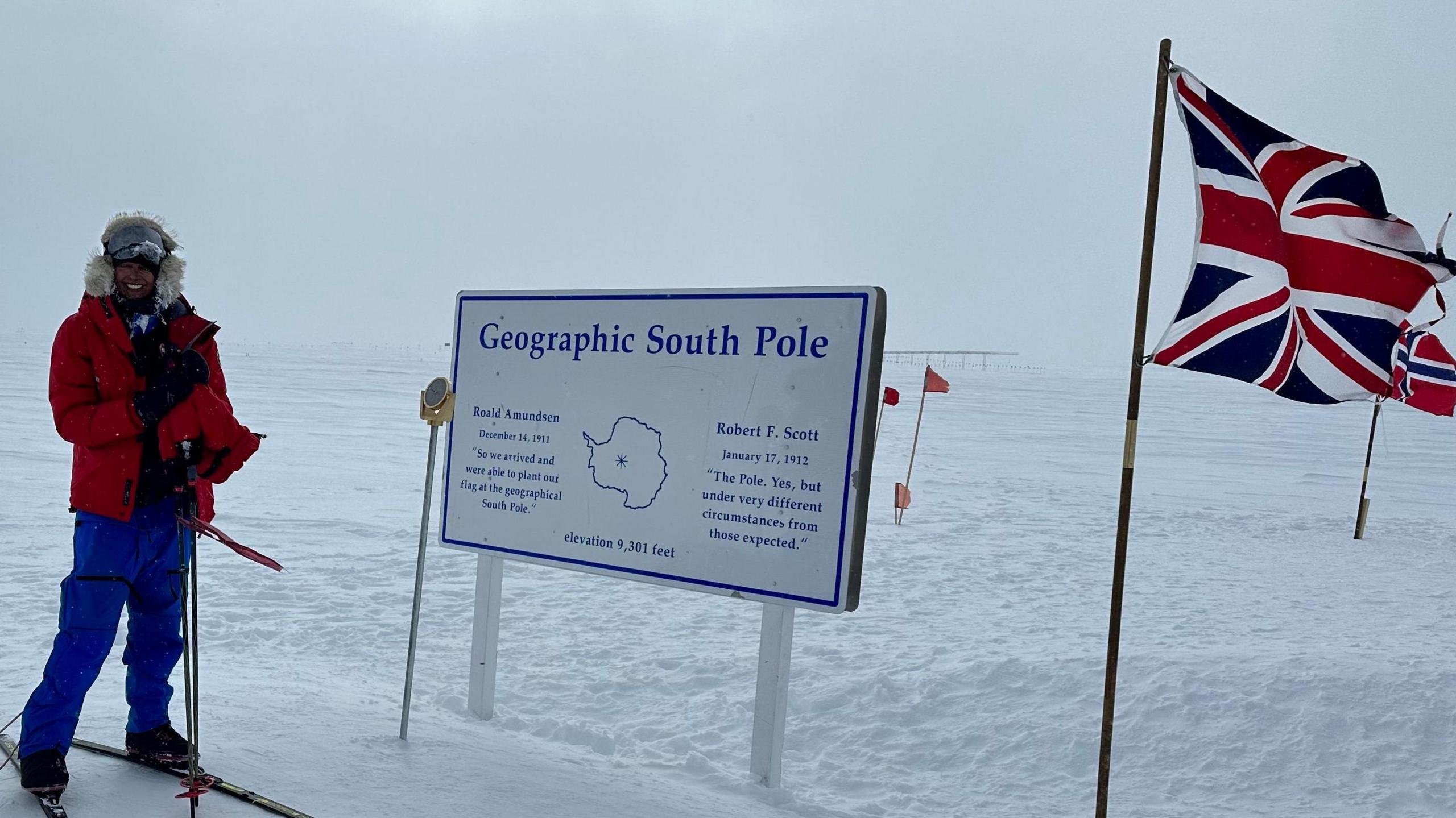 Harpreet Chandi is pictured wearing a red coat, blue trousers and skis. SHe is smiling next to a sign which says Geographic South Pole in the middle and the Union flag to the right.