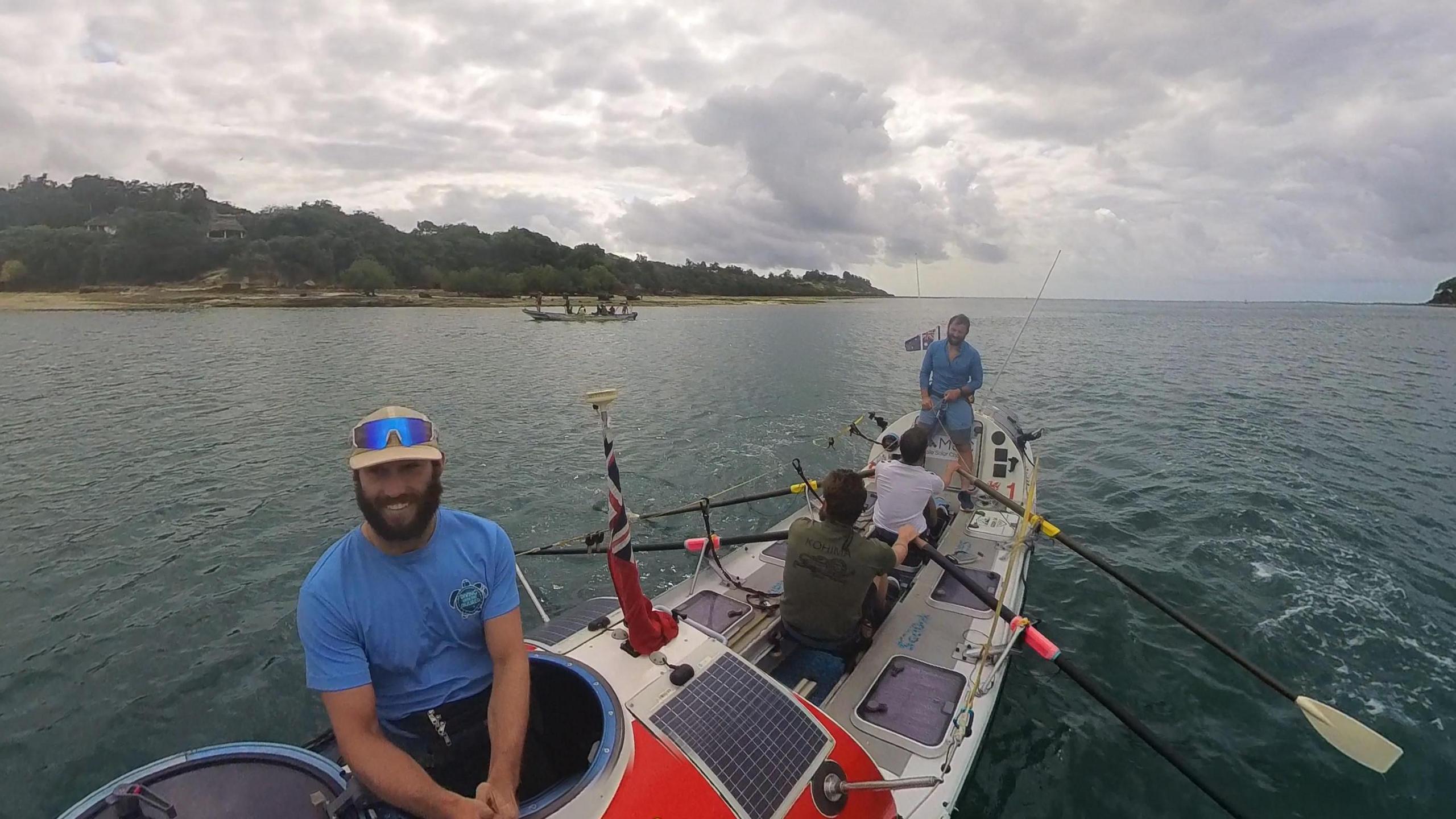 Four men on a large white rowing boat. One is sitting in the cabin on the left and smiling at the camera. Two of them are rowing and one is sitting at the front directing them. Behind them is an ocean and some land in the distance.