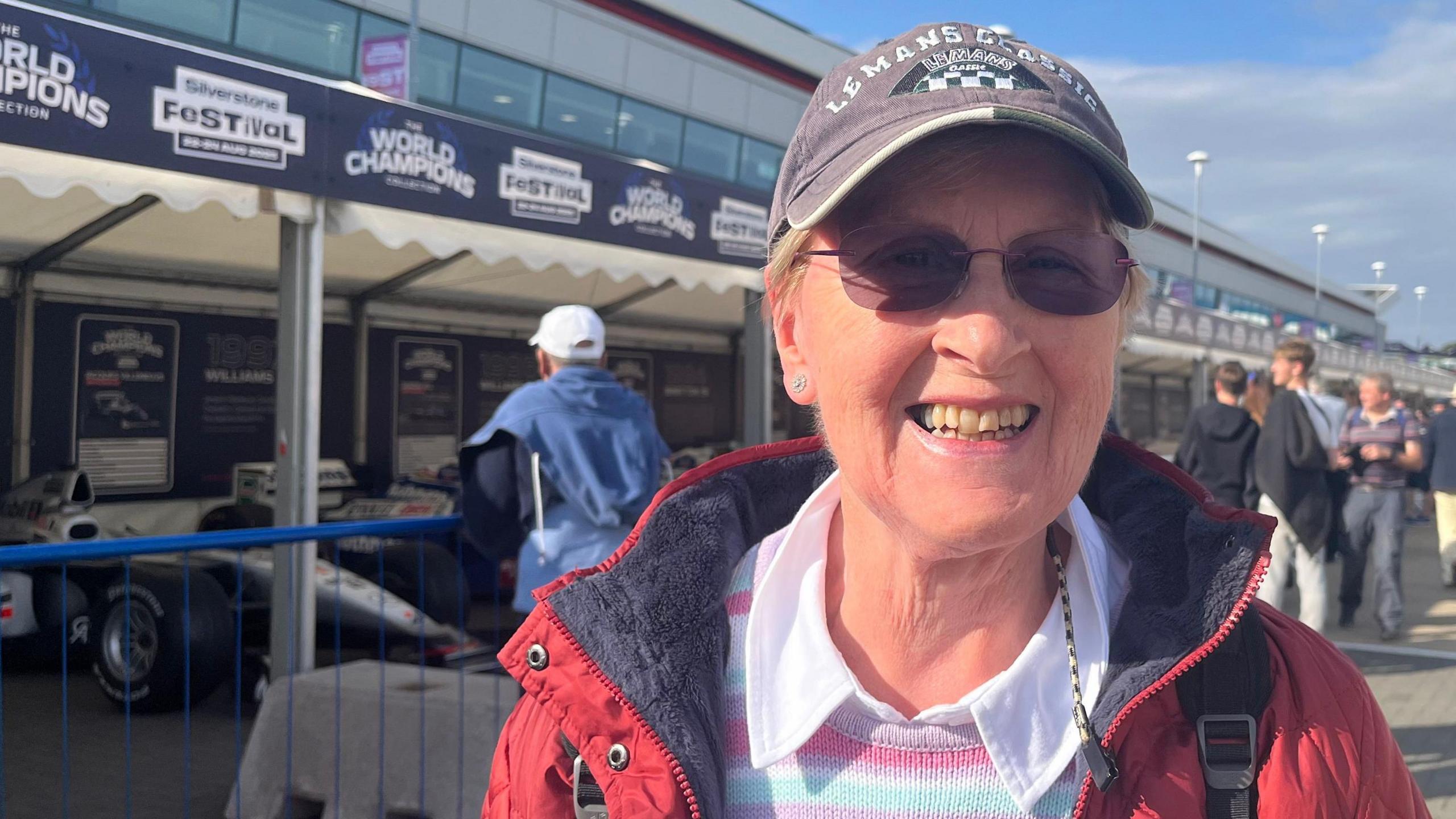 A woman wearing a red coat, sunglasses and a Le Mans cap is smiling at the camera. She is standing in front of the World Champions sign and there are groups of people lining up to look at the cars.