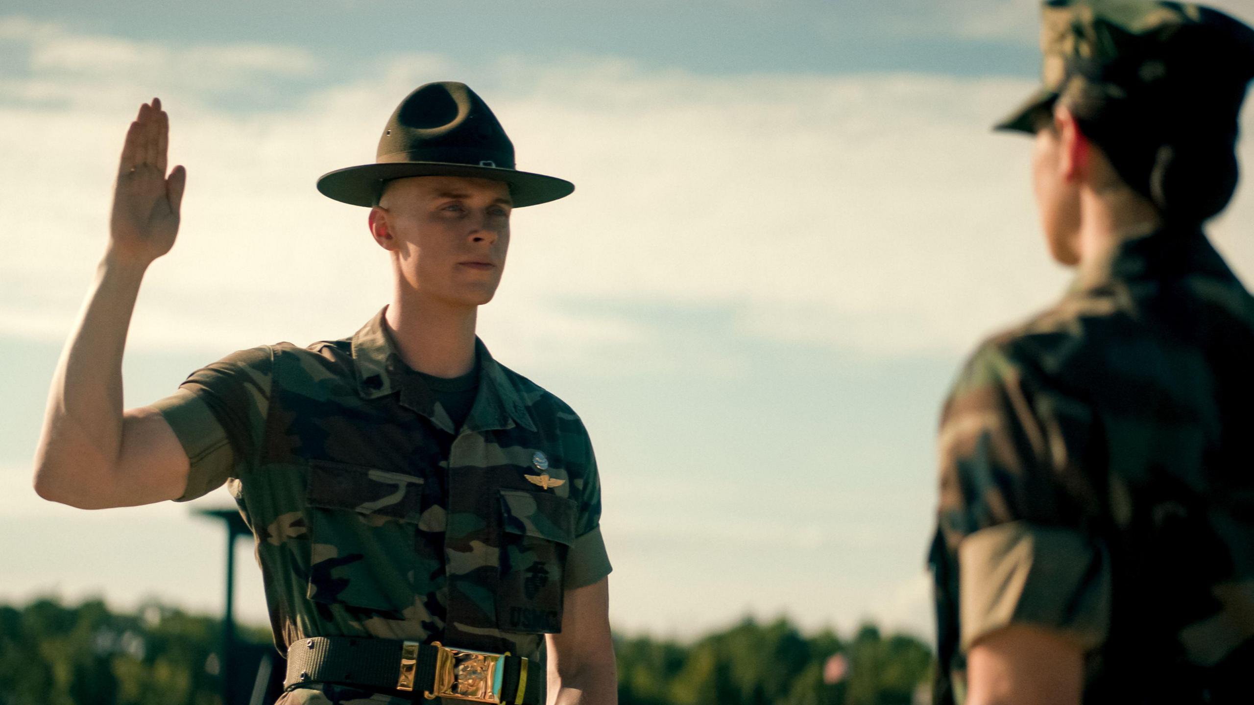 Max Parker as Sgt Sullivan, wearing a camouflage green military shirt and green brimmed uniform hat. He raises his hand straight as if swearing an oath in front of a female superior, who is also wearing a camouflage military shirt and cap. They are outdoors with trees behind.