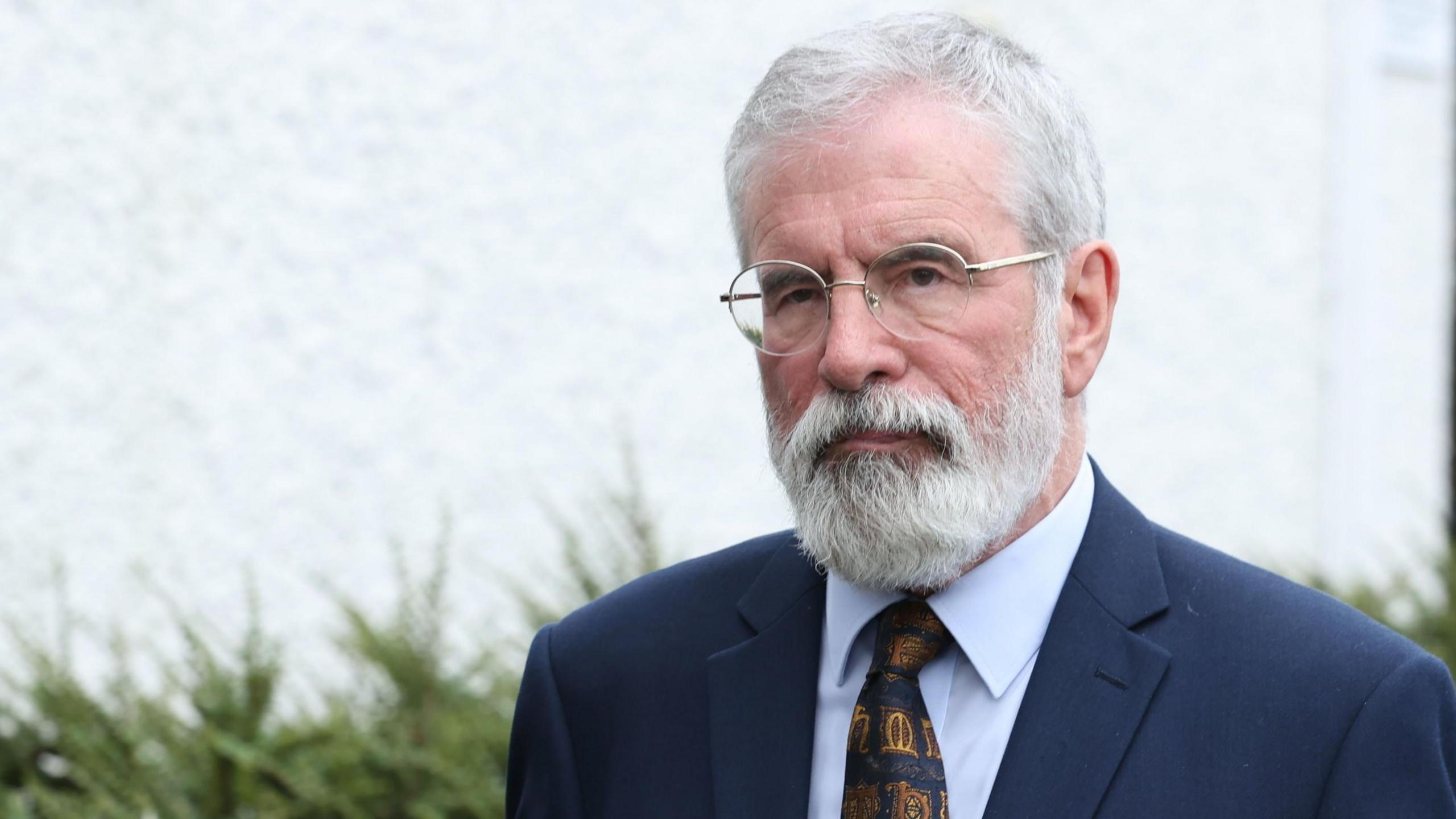 Gerry Adams has a white beard and wears glasses. He's wearing a navy suit with a blue shirt and a tie.