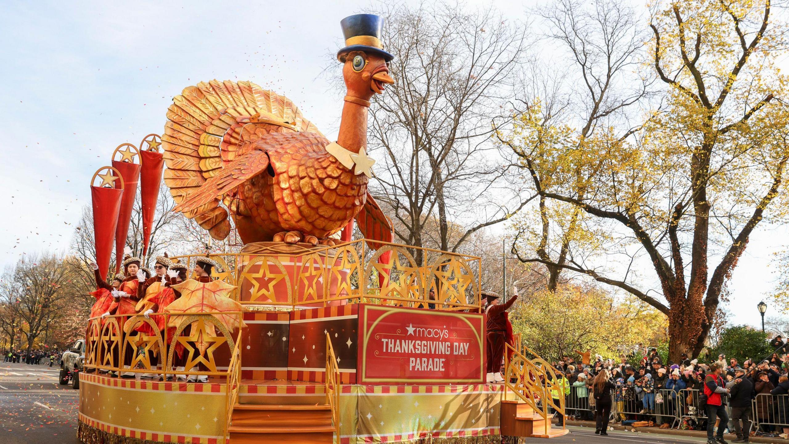 A huge parade float of a turkey wearing a pilgrims hat. Everything is golden orange coloured.