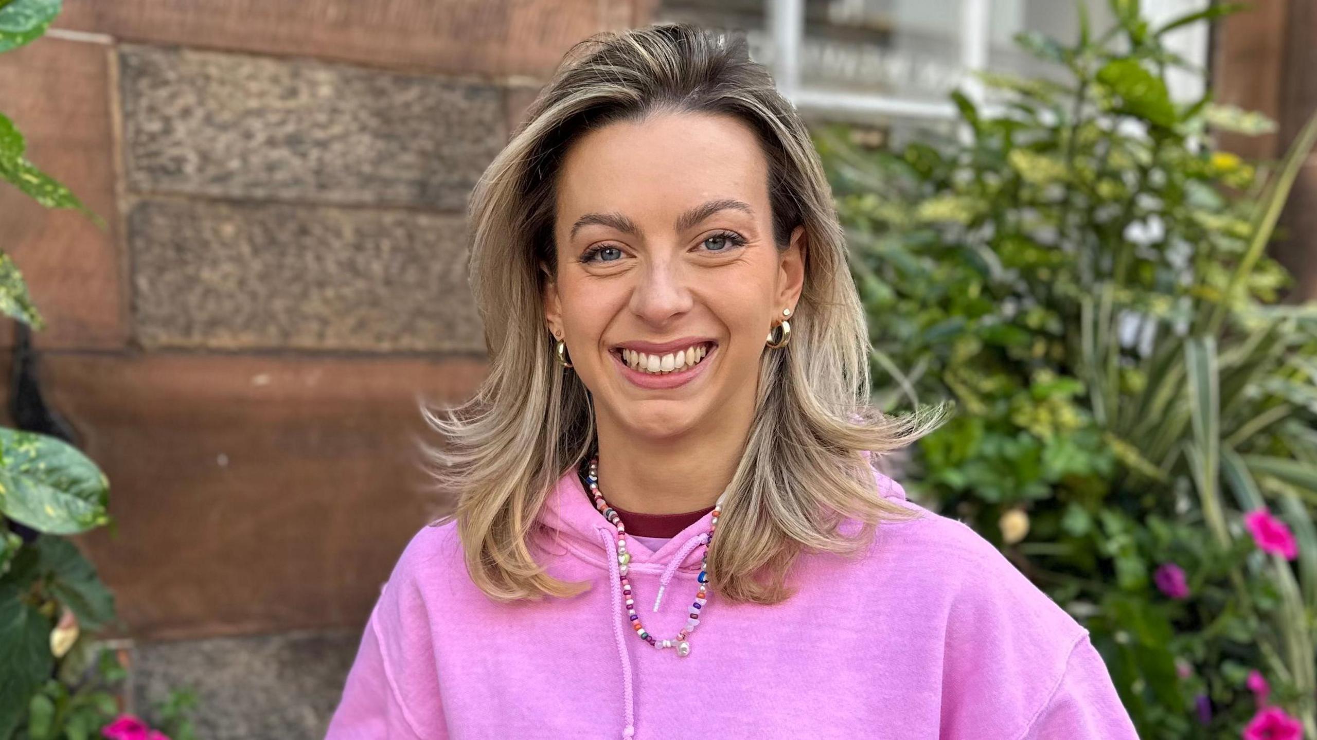 Abi Clarke. Abi's a white woman in her 20s with shoulder length blonde hair and blue eyes. She smiles at the camera, wearing a pink hoodie, gold hooped earrings and a beaded necklace. She's pictured outside against a brick wall with flowers and plants around her. 