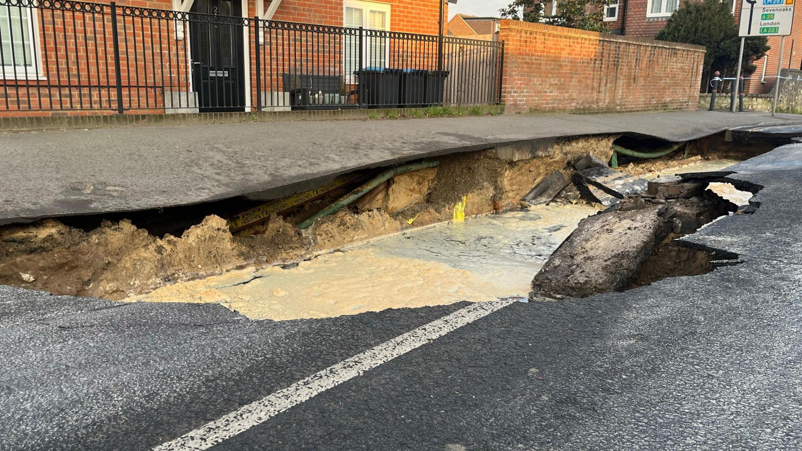 A view of the sinkhole taken from the level of the road surface. It shows the ground beneath the pavement, now exposed by the sinkhole that has opened up in the road. Pooled water is visible inside the hole. A red brick house with black metal fencing sits behind the pavement.