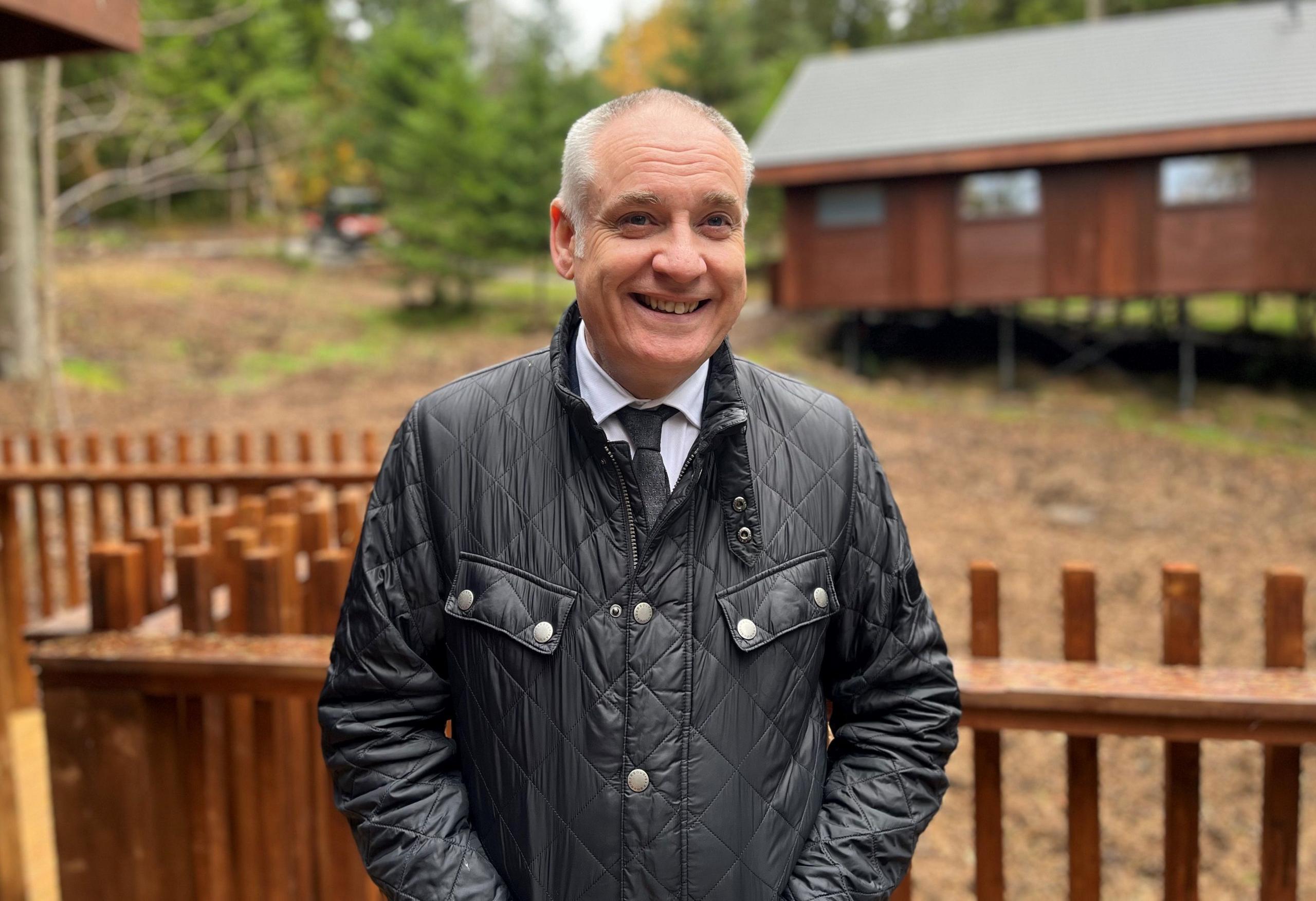 Minister for business Richard Lochhead smiling at the camera as he stands outside a new timber cabin at Glentress