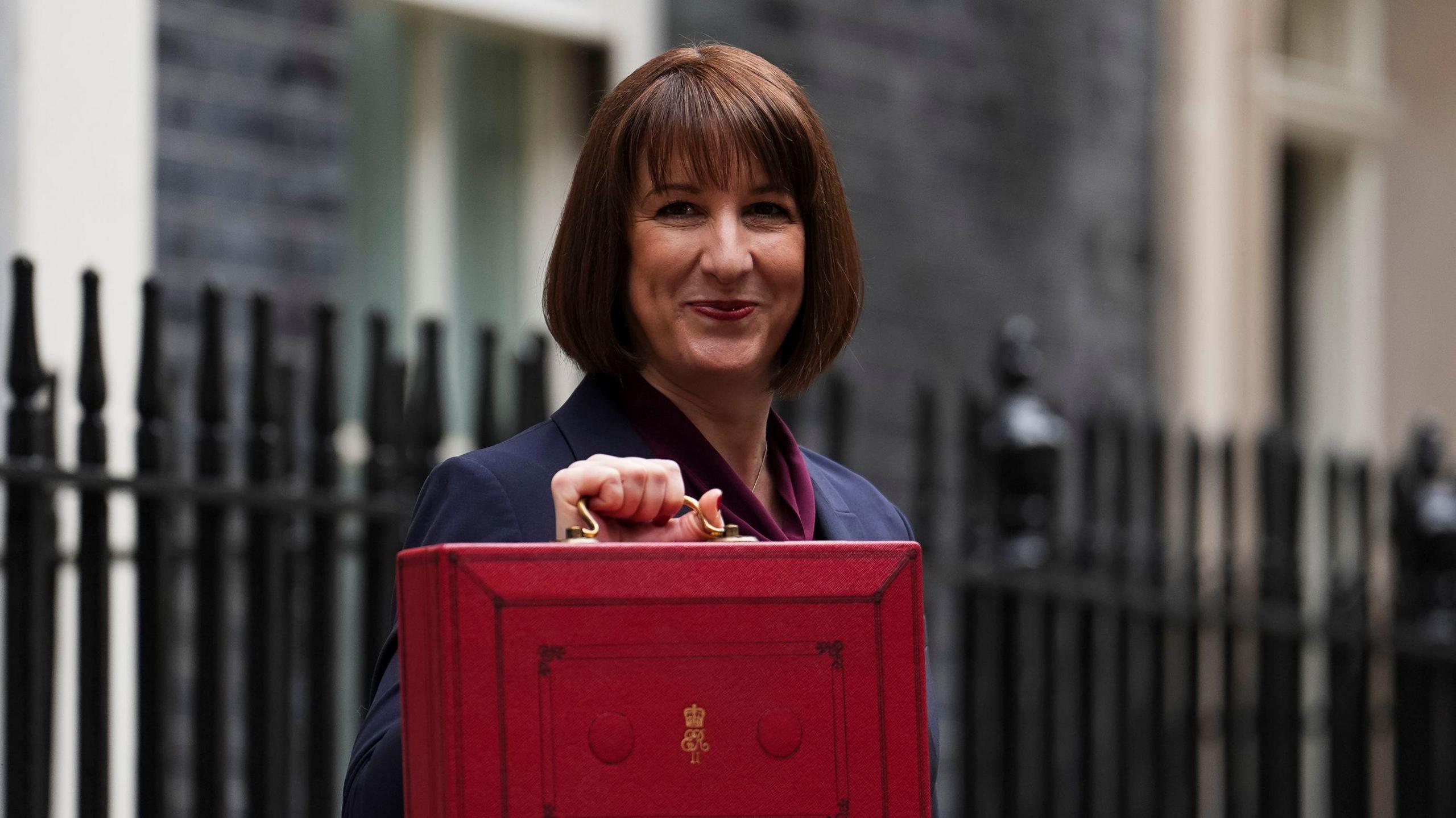 Rachel Reeves dressed in a blue suit and a maroon scarf holding a red briefcase. Behind her are black metal railings and behind them the blurred sides of buildings.