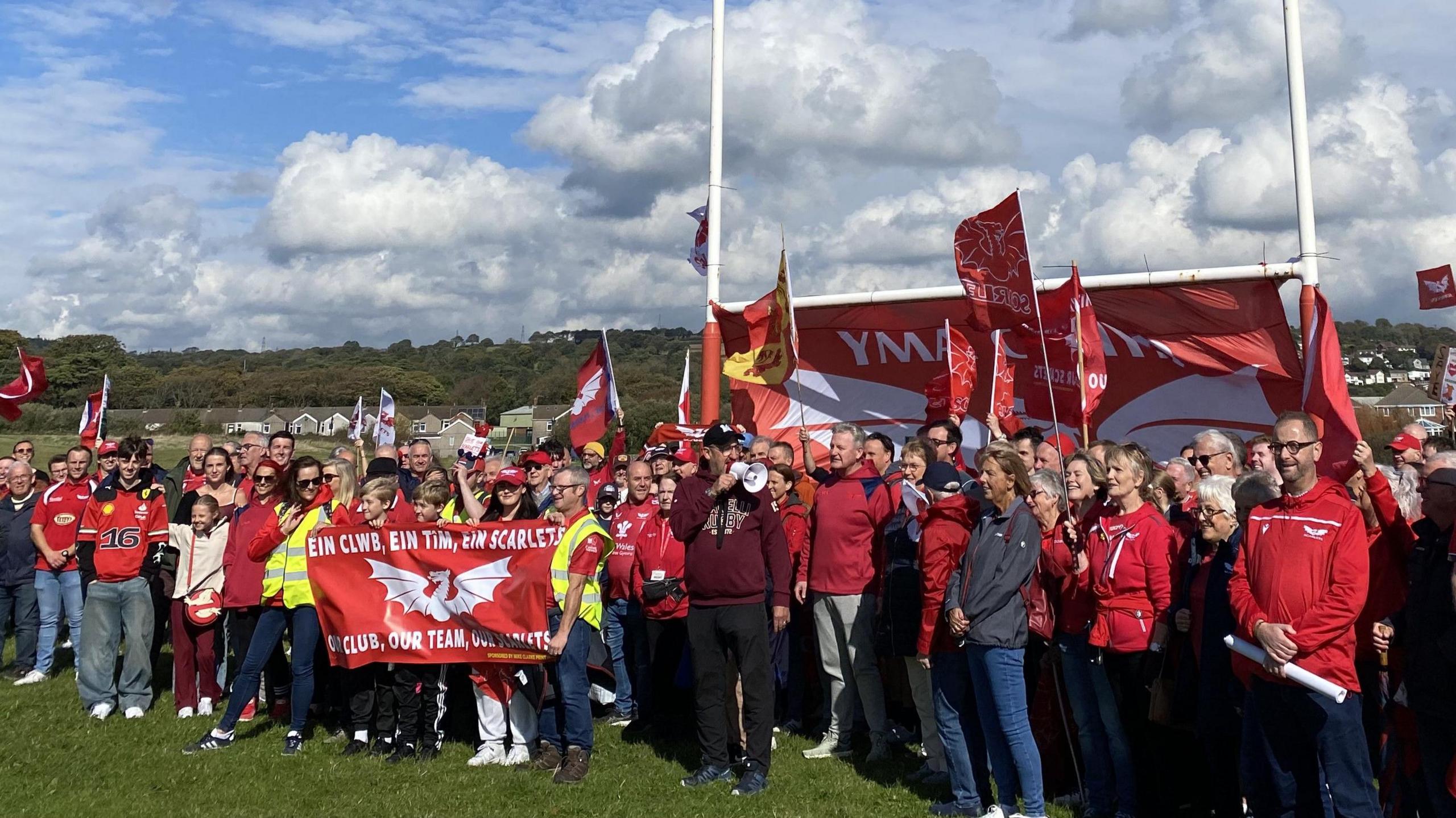 A large group of a peple are gathered on a rugby pitch beneath the goal posts. Most are dressed in Scarlets red jerseys and some are holding flags 
