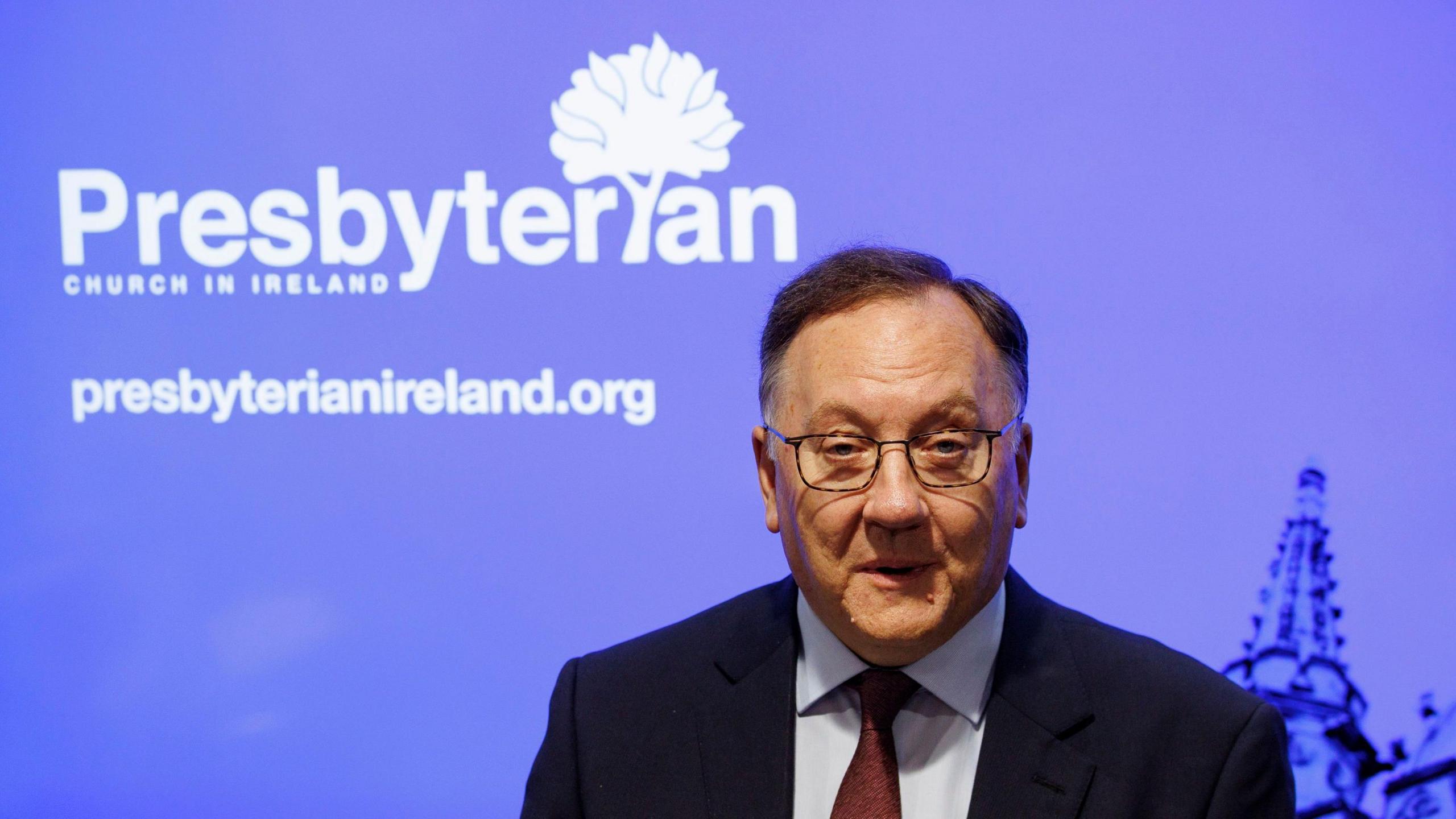 Dr Gribben has short dark hair combed to one side. He is wearing glasses and dressed in a black jacket, light blue shirt and a red tie. Behind him is a backdrop with the image of church spire and the words Presbyterian Church in Ireland.