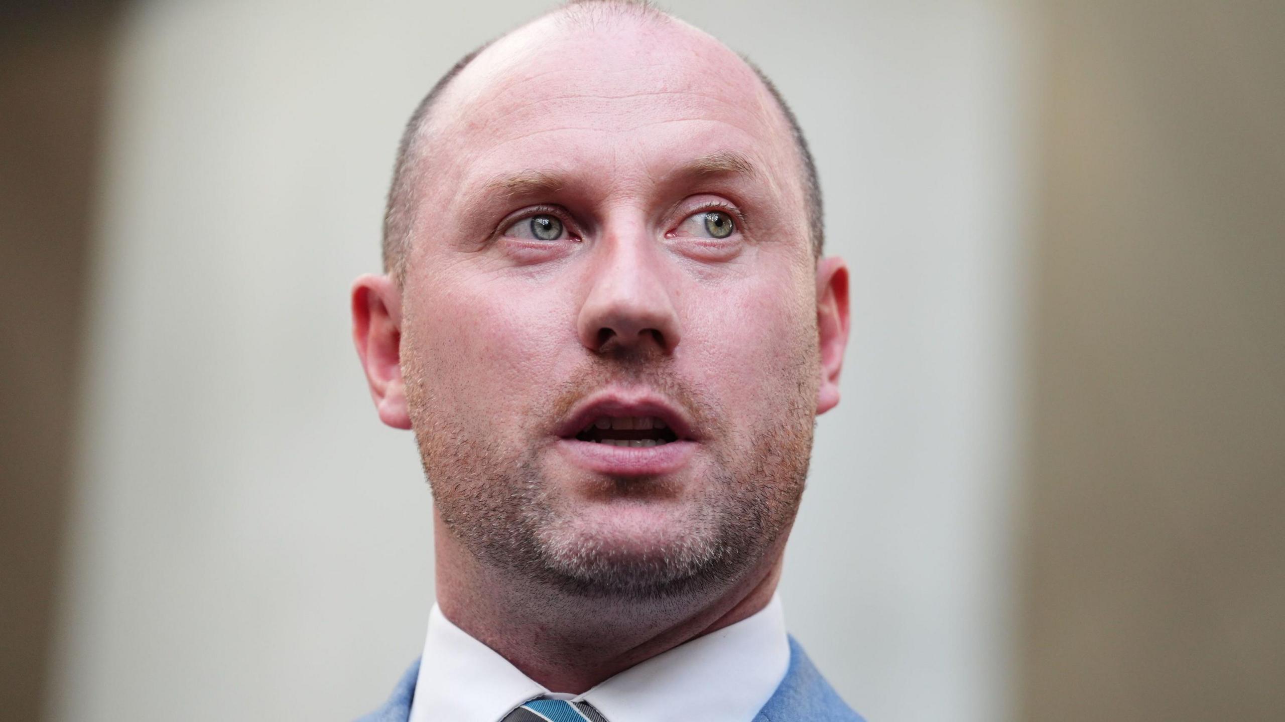 A man wearing a light blue suit and tie. He is balding, with a goatee (or possibly just unshaven) The background is out of focus.
