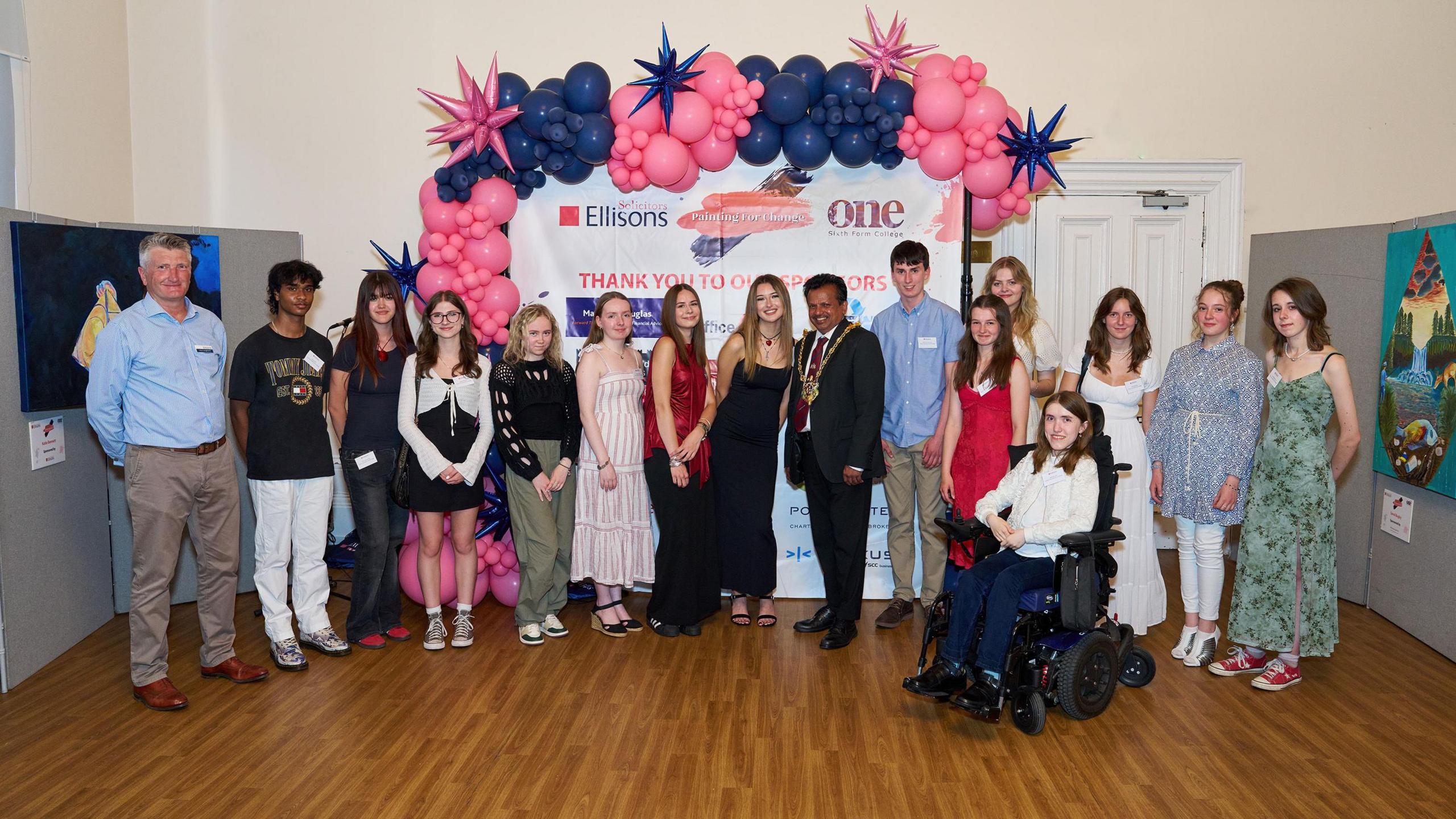 Art students from One Sixth Form College standing in a line and looking directly at the camera, with the mayor of Ipswich dressed in a suit and wearing a mayoral chain. Behind them are balloons and a screen reading "Thank you to our sponsors". 