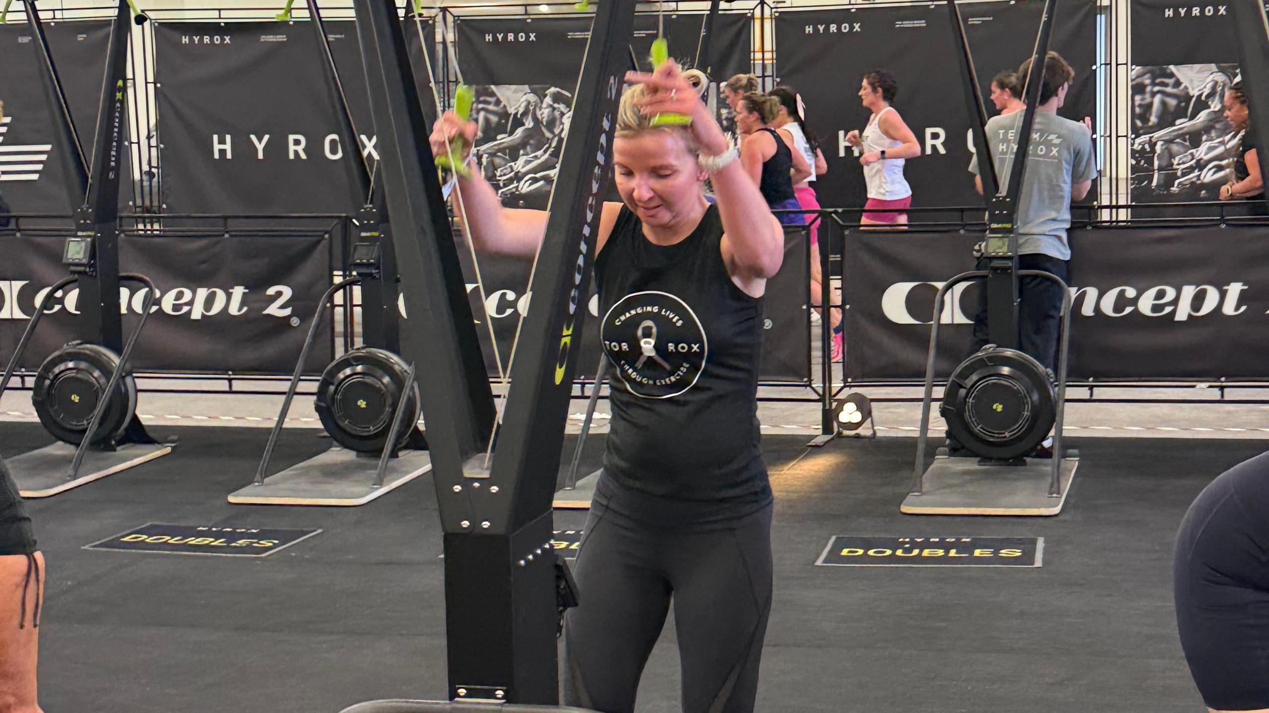 Tor competing in a Hyrox competition, she is on a gym machine, with people behind her. She is wearing a black top and bottoms with her hair tied up.