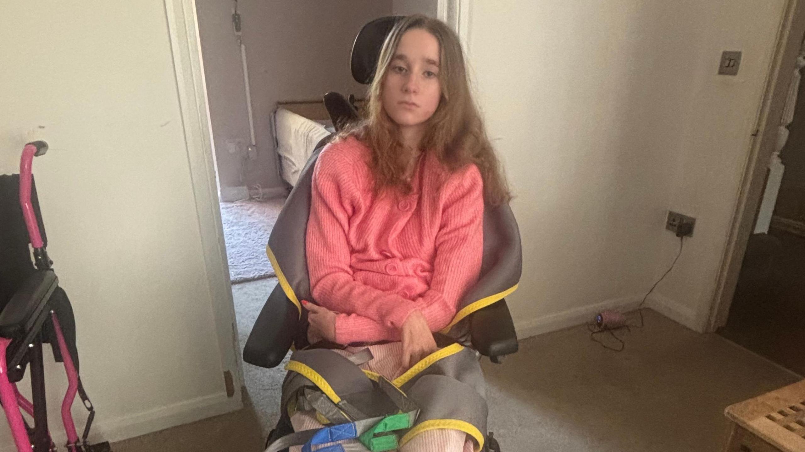 A young girl with brown hair in a wheelchair. She is wearing a pink top and has her arms crossed in front of her. There is a pink wheelchair slightly visible on the left side of the image.