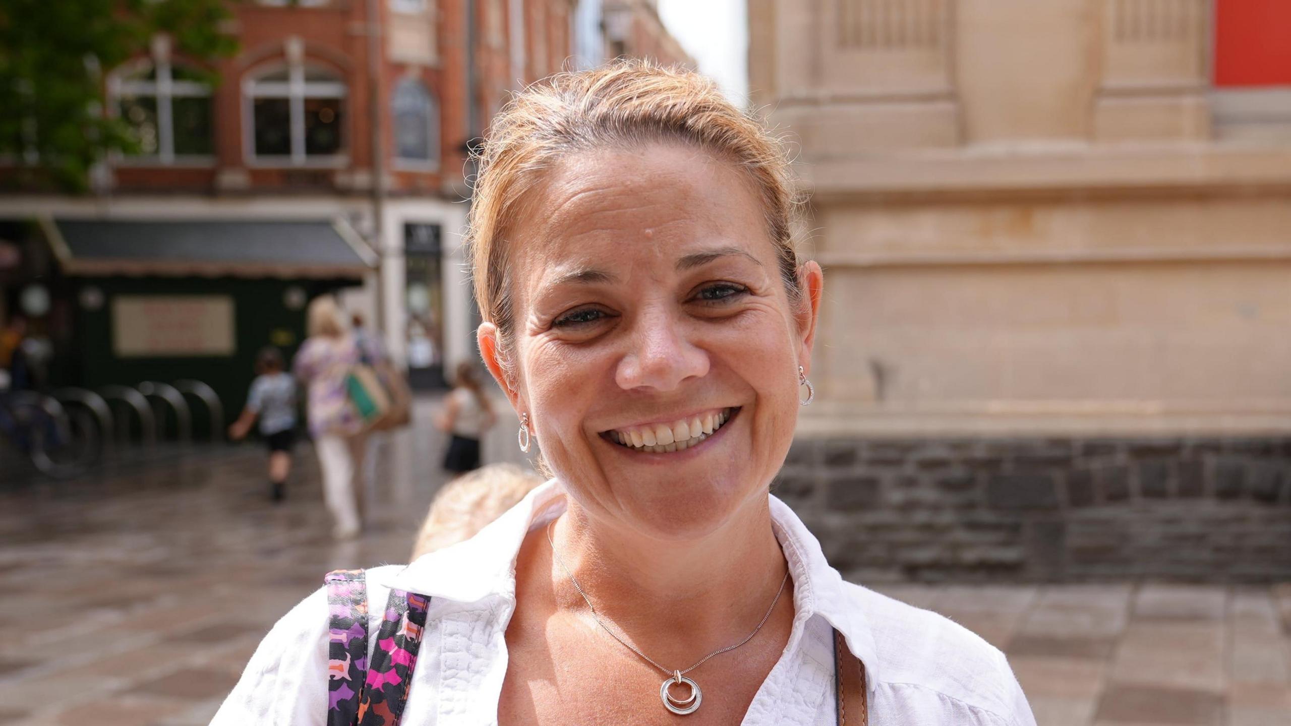 Jane Hides has her blond hair pulled into a ponytail and is wearing a matching circular set of earrings and necklace. She is wearing a white collared shirt