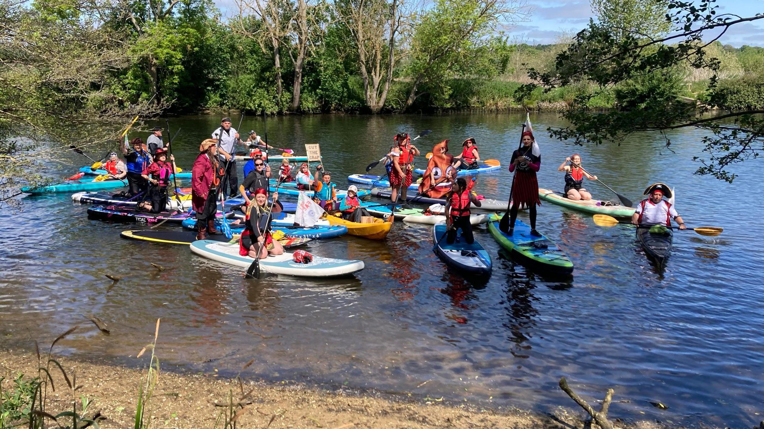 About 20 people stand or sit on paddleboards and canoes near the bank of the river, posing for a photograph. It is a sunny day and many are wearing bright colours and pirate hats/costumes.