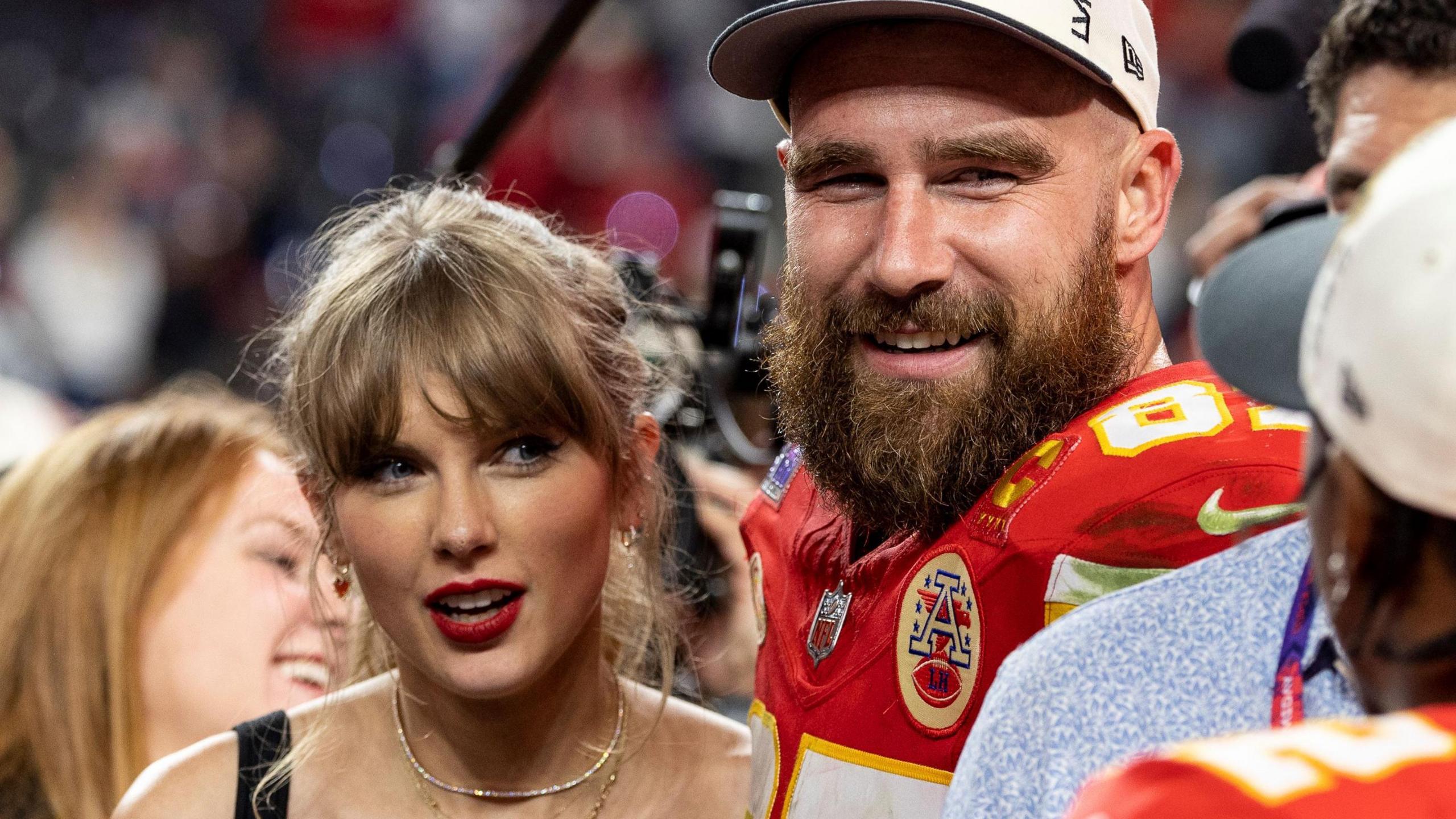 Taylor Swift and Travis Kelce after the Kansas City Giants won the Superbowl. Taylor wears her blonde hair tied back and a red lipstick. Travis wears his red Kansas kit with a white cap. He has a thick beard and smiles. They're surrounded by photographers and fans. 