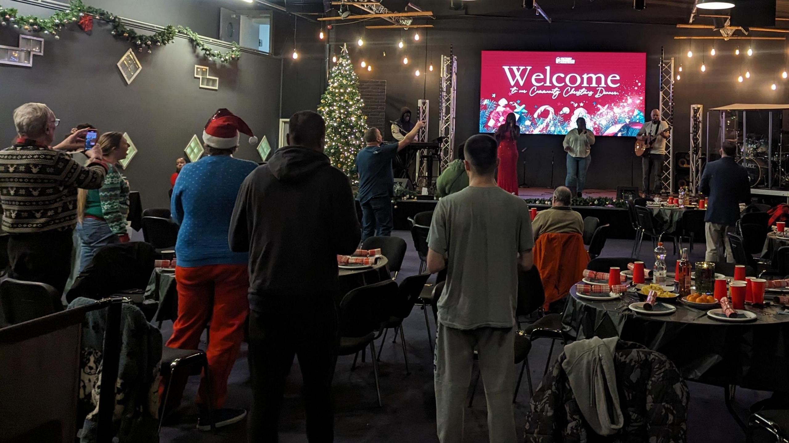 Backs of guests, who are standing by tables with plates and crackers in a large room, as they watch singers and musicians perform on stage in front of a large screen saying "Welcome"