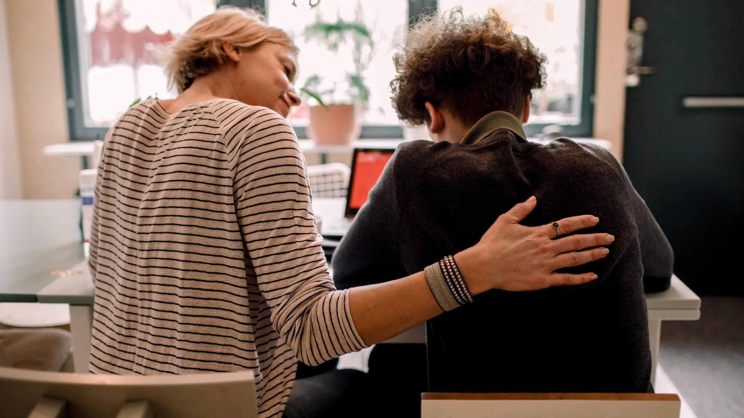Stock photo of mother, wearing a stripy top, with short blonde hair, sitting by teenage son, with brown curly hair, studying at home - at a table, in front of a laptop.