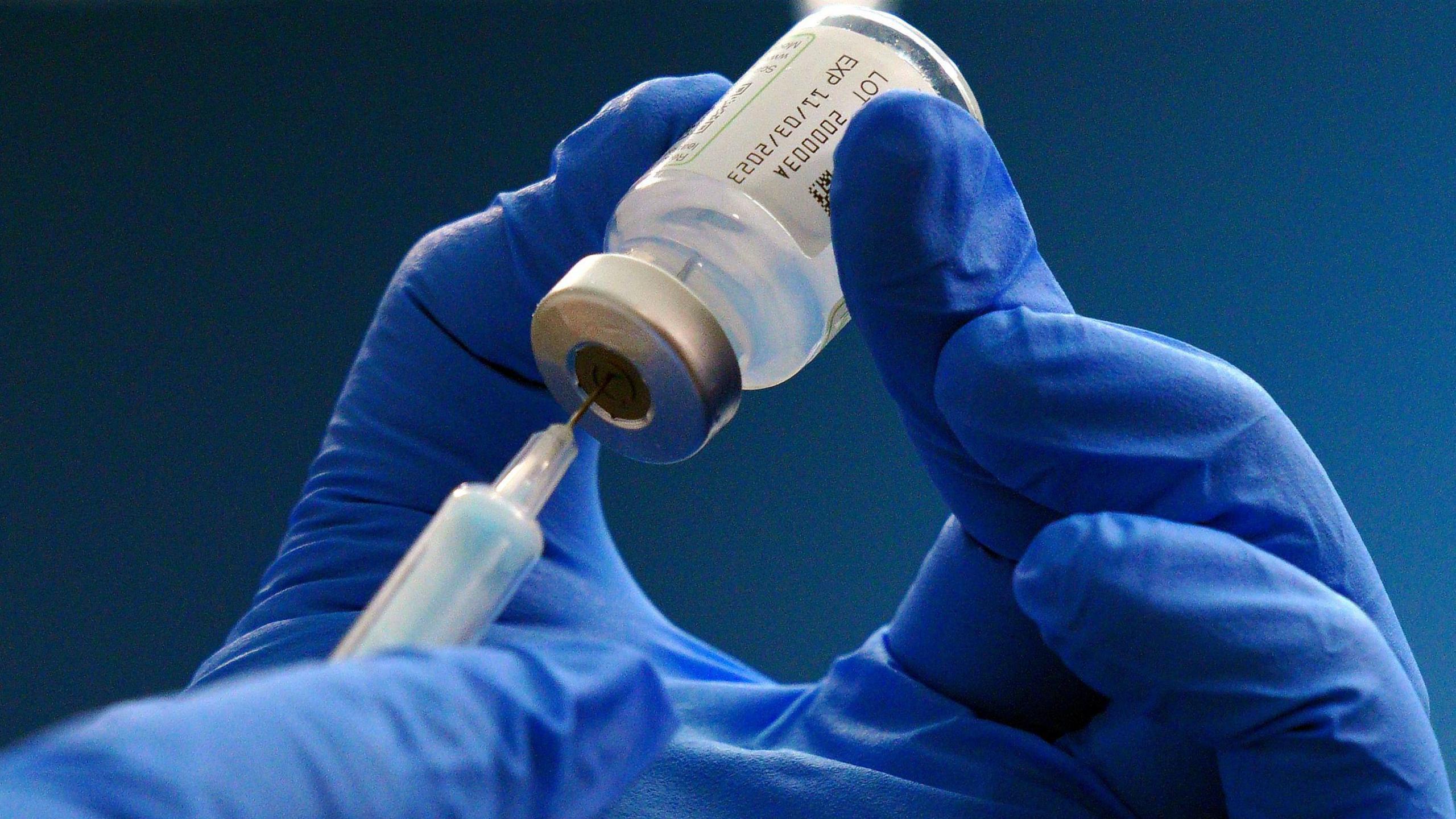 A pair of hands with blue gloves on holding a syringe extracting medicine out of a vial.