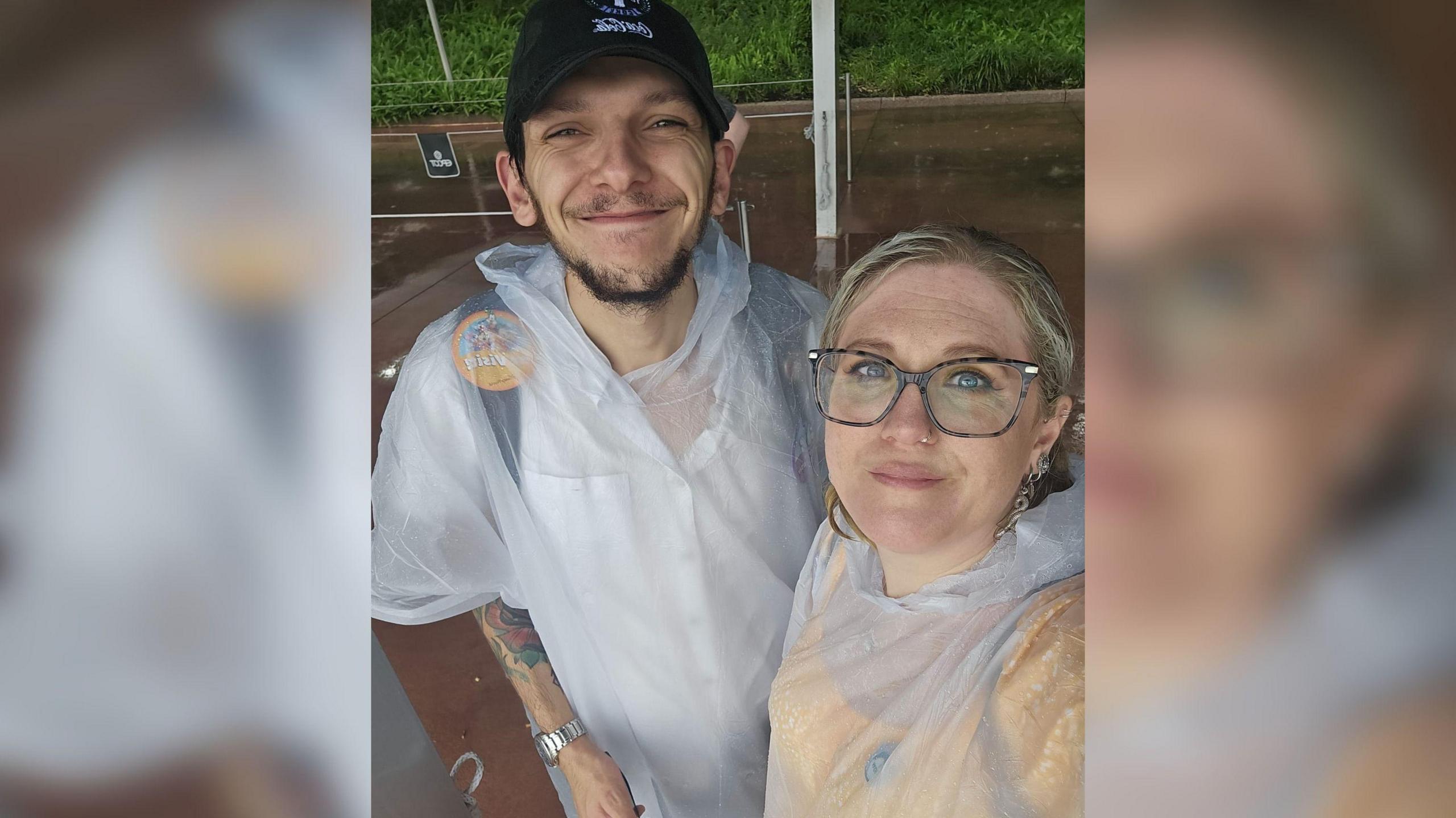 A selfie of a man and woman who are wearing clear ponchos. The man is wearing a black cap and the woman has glasses on