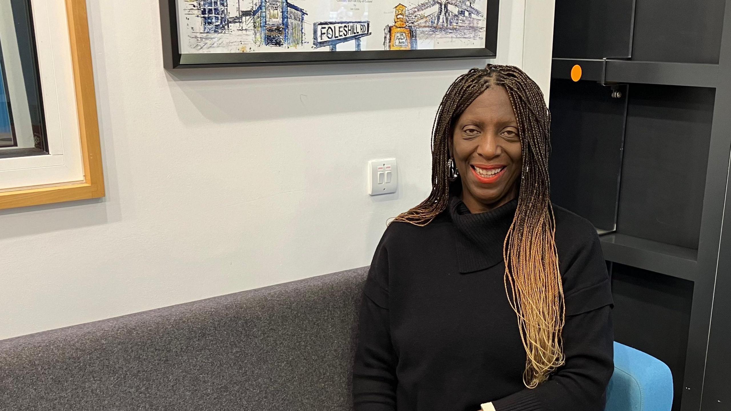 A woman smiling at the camera while sat on a sofa. She is wearing a black polo neck top and has long dreadlocked hair.