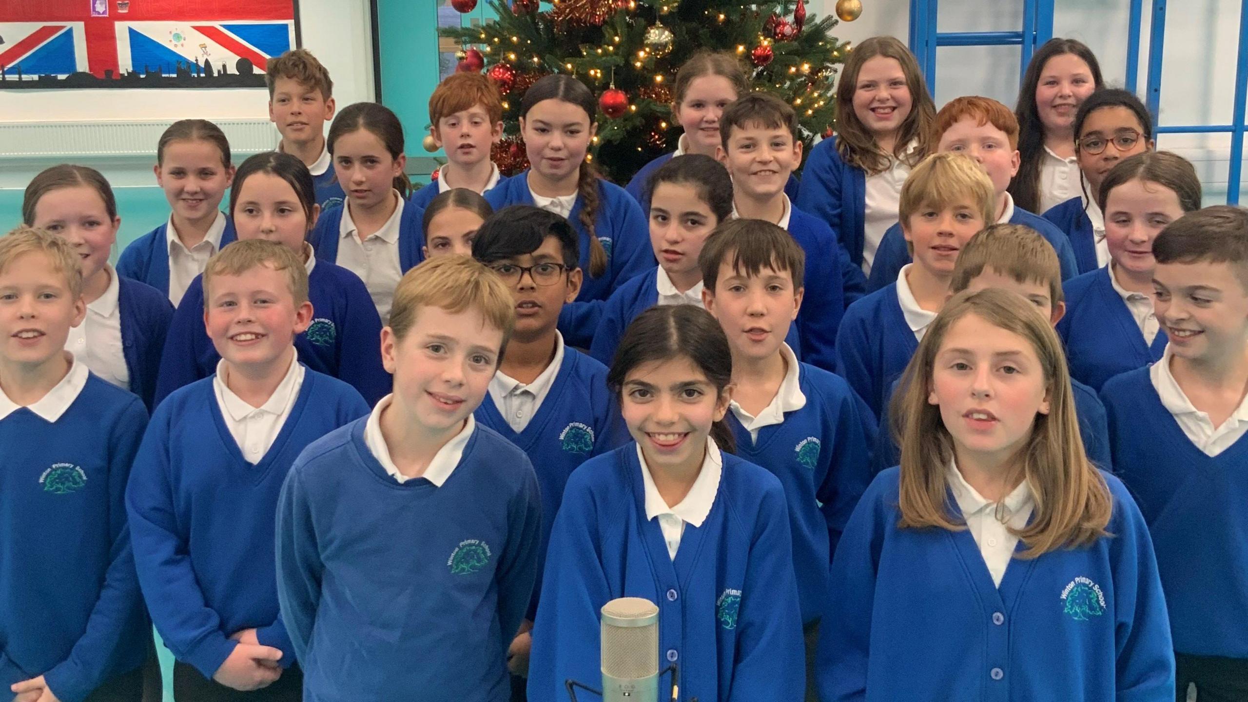 A group of children in blue and white school uniform looking at the camera, with a microphone in front of them and a Christmas tree behind them.