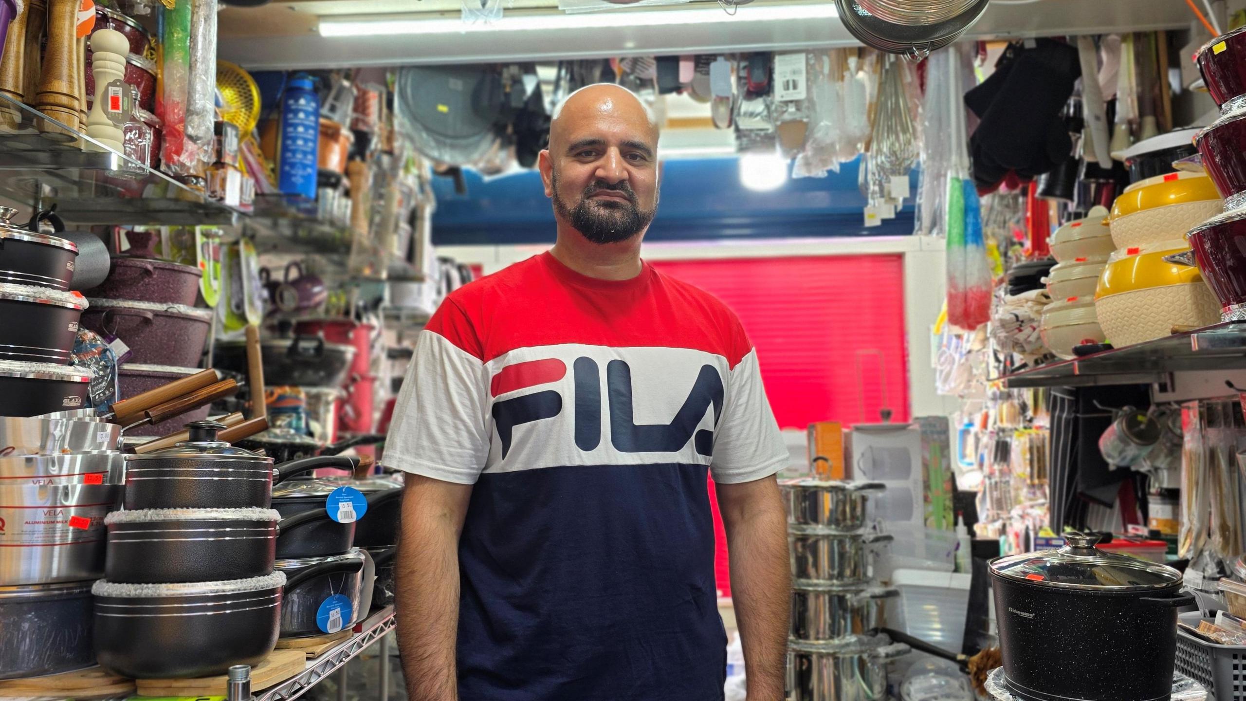 Mohammed is wearing a blue, white and red 'Fila' t-shirt. He's standing in the middle of the stall, surrounded by cookware.