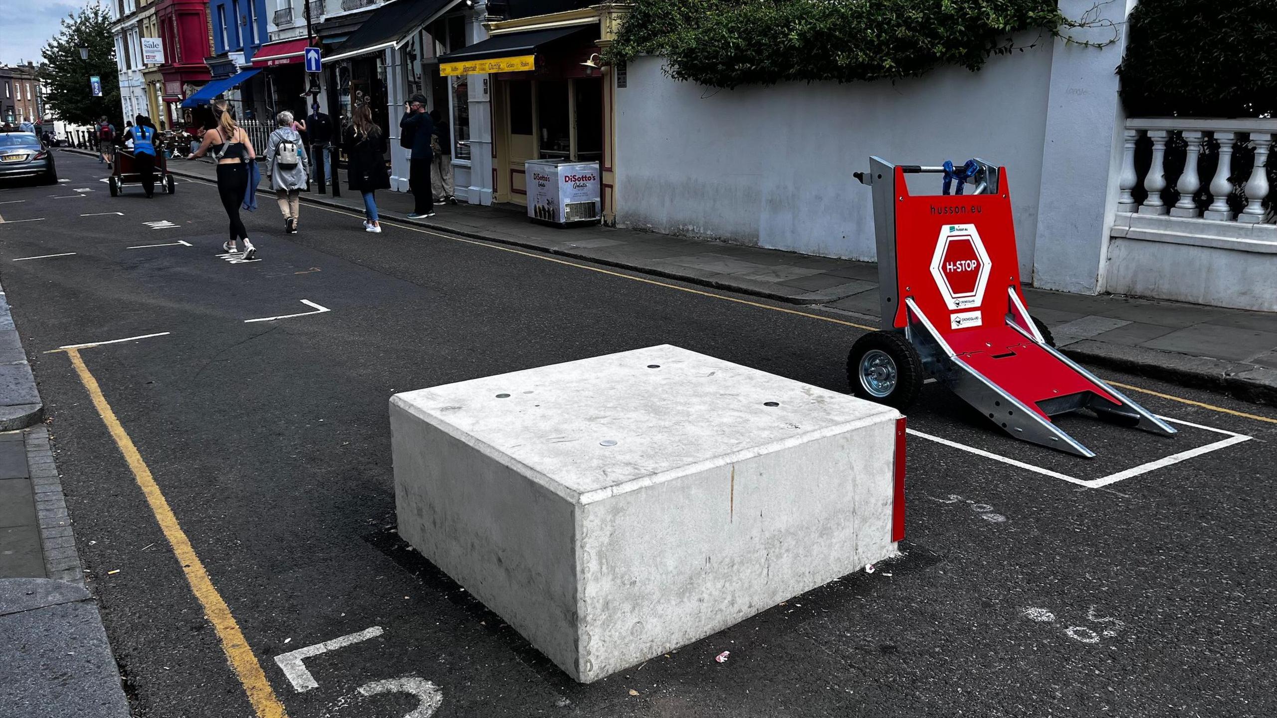 A flat, light grey concrete block and a large red clamp on wheels are blocking a tarmac road. The road has a few white markings denoting parking spaces, and a single yellow line running along both sides. There is a narrow pavement on both sides of the road, with the photo taken from the pavement on one side looking across and down the street. A few people are walking in the road in the background, and there are some shops in the background lining the street.