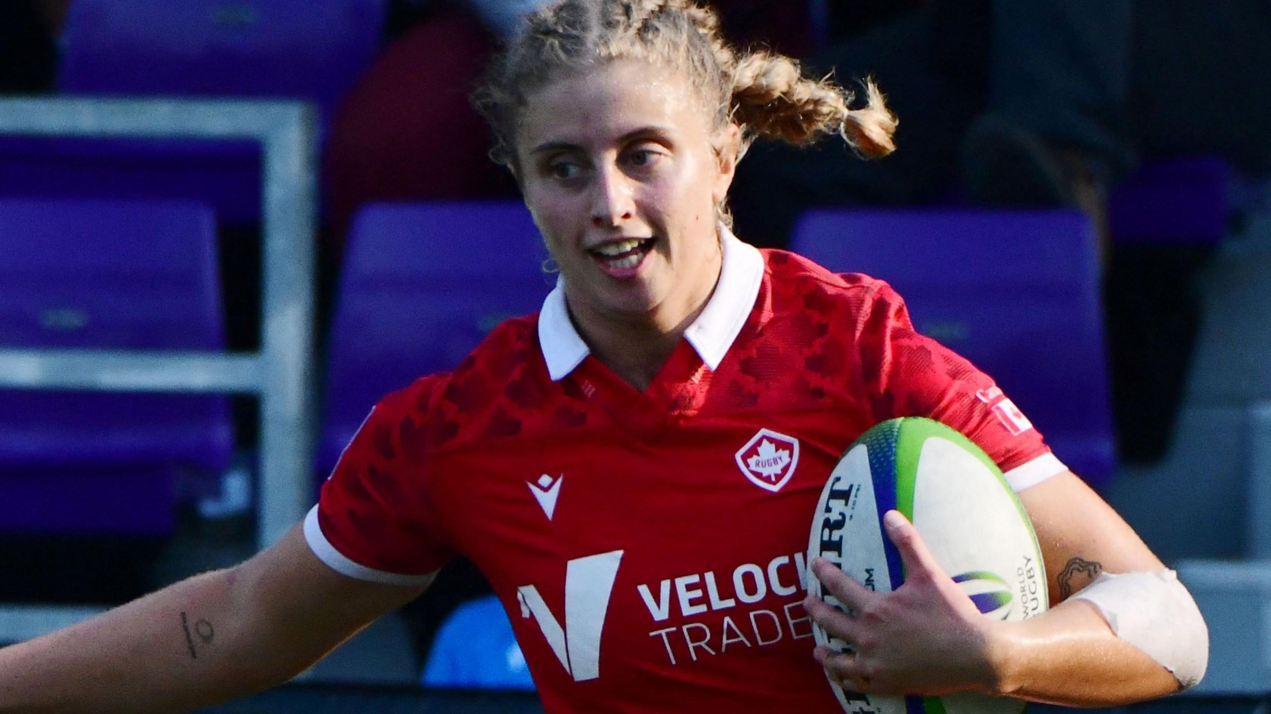 Maddy Grant in action for Canada, sporting a braided light-haired ponytail, wearing a red shirt, cradling a rugby ball with her left hand and arm