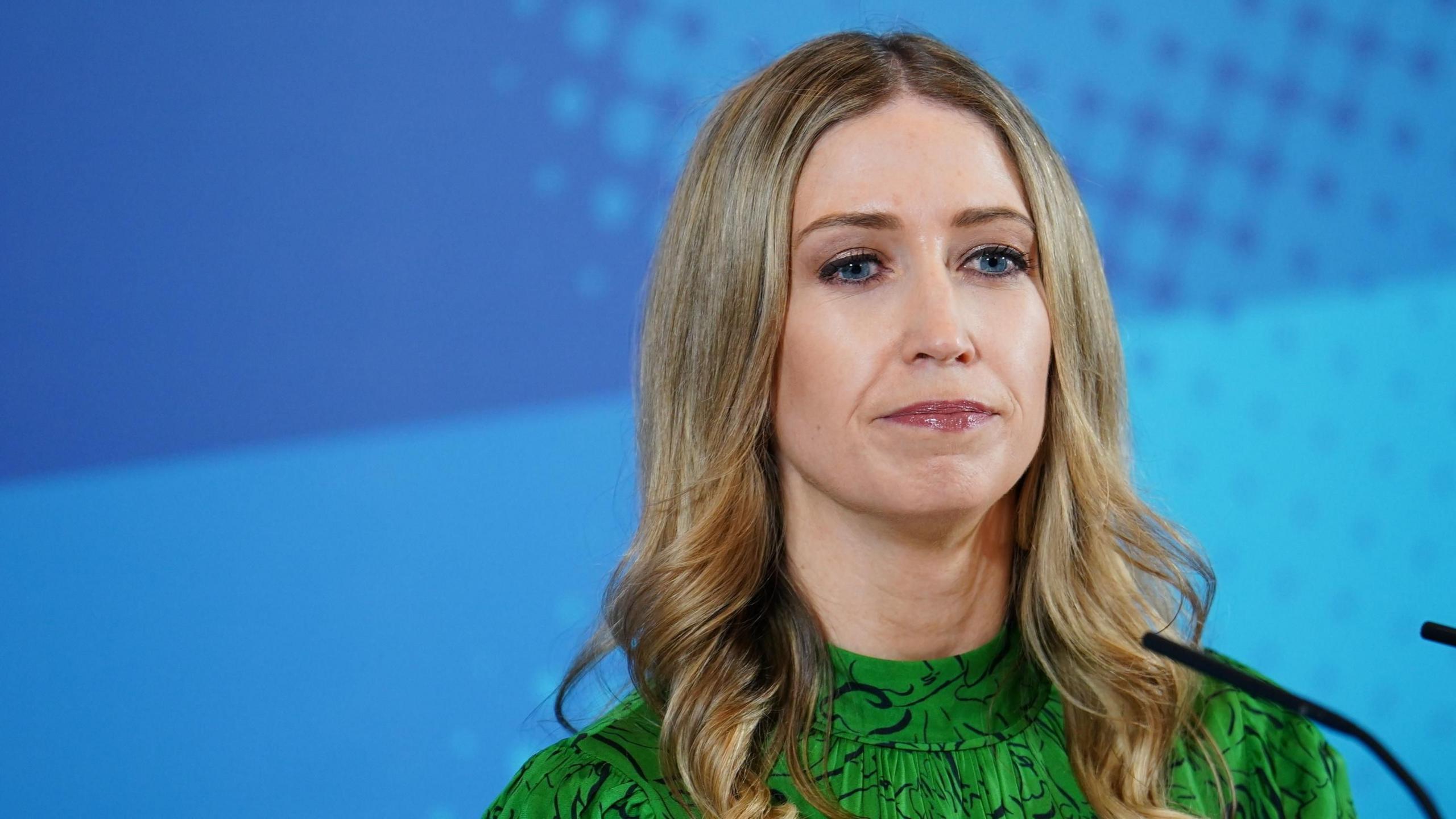 The head and shoulders of Laura Trott MP. She is wearing a green dress and stands against a textured blue background. She is framed to the right, with a microphone entering the shot in the bottom right hand corner.