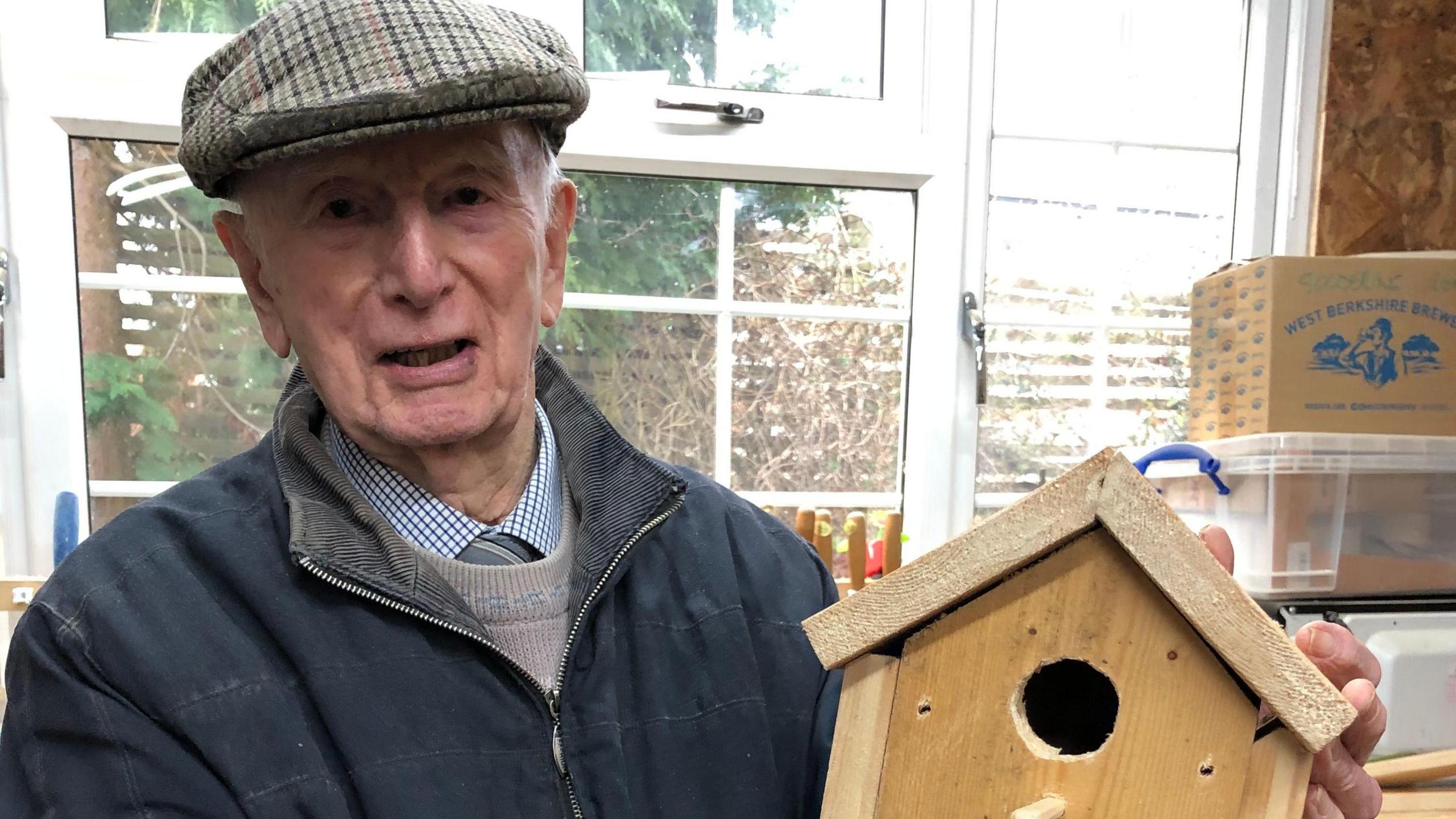 Ray Frewing wears a flat cap and a dark blue zip-front jacket with a beige jumper over a blue and white gingham shift and a green striped tie. He is holding a wooden birdbox as he stands in front of a window through which the sun is shining.