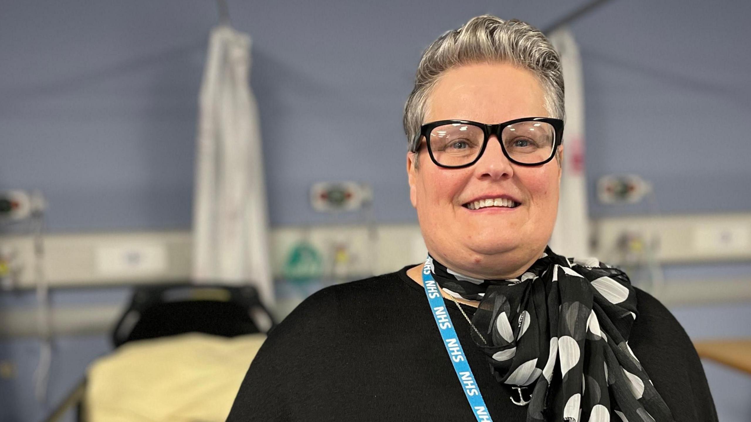 Rachael Hardcastle-Pearce, a woman with swept-back silver hair and black-rimmed glasses, smiles as she stands in a hospital ward. She is wearing a black top, black and white scarf and a blue NHS lanyard.