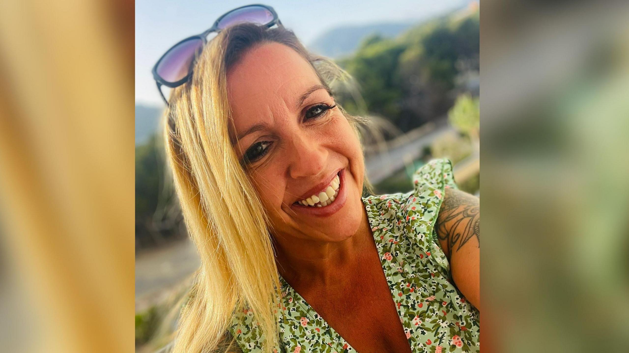 Ali Alcock smiles at the camera in this selfie. She has long blonde hair with sunglasses on the top of her head. She is wearing a light green top with a pink floral design. Blurred trees can be seen in the background.