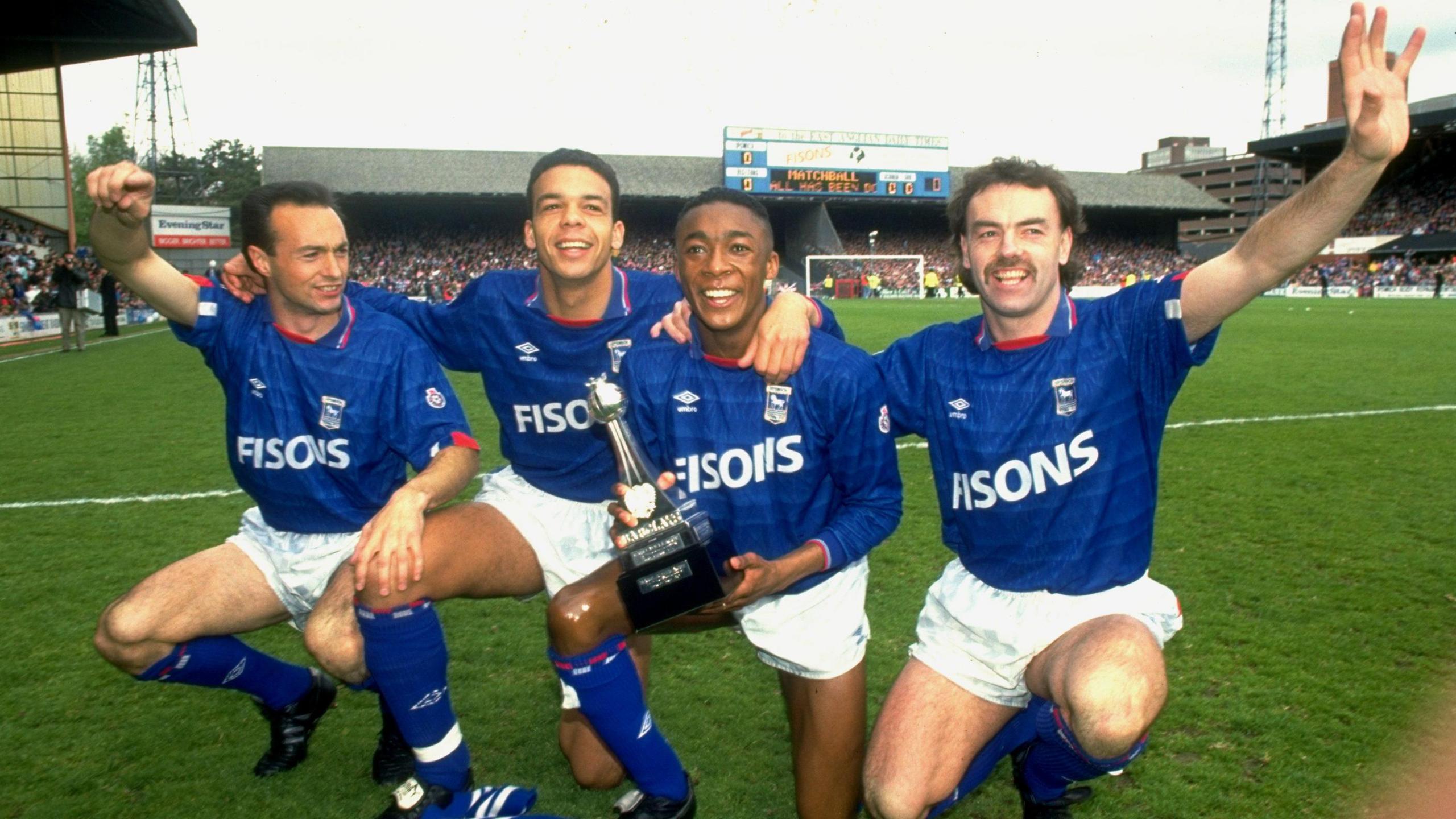 Simon Milton, Jason Dozzell, Chris Kiwomya and John Wark crouching on the pitch at Portman Road in their Ipswich Town kits, with the old Second Division trophy being held by Kiwomya.