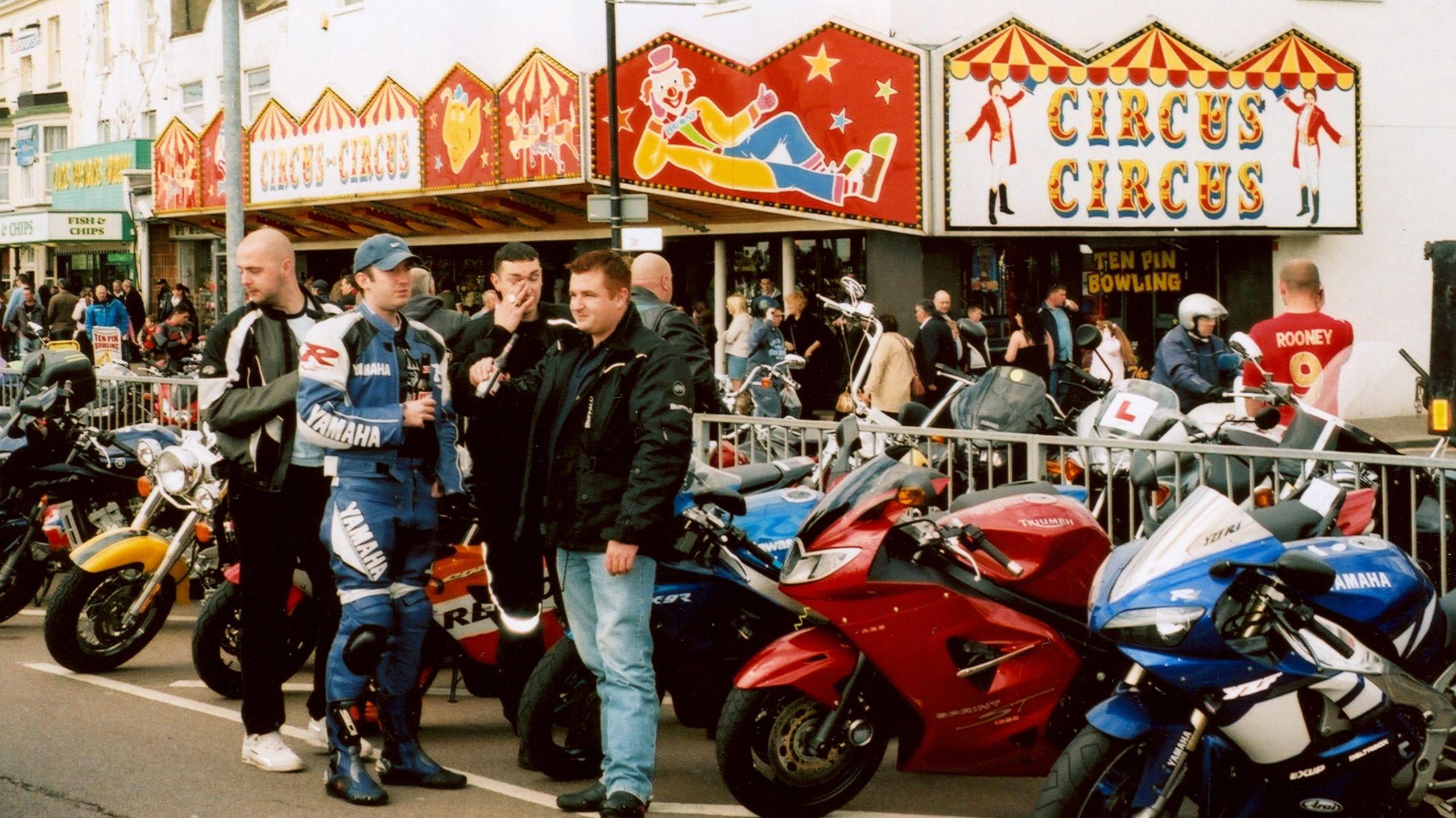 A group of men standing next to their bikes. Behind them in a circus building.
There is quite a big crowd