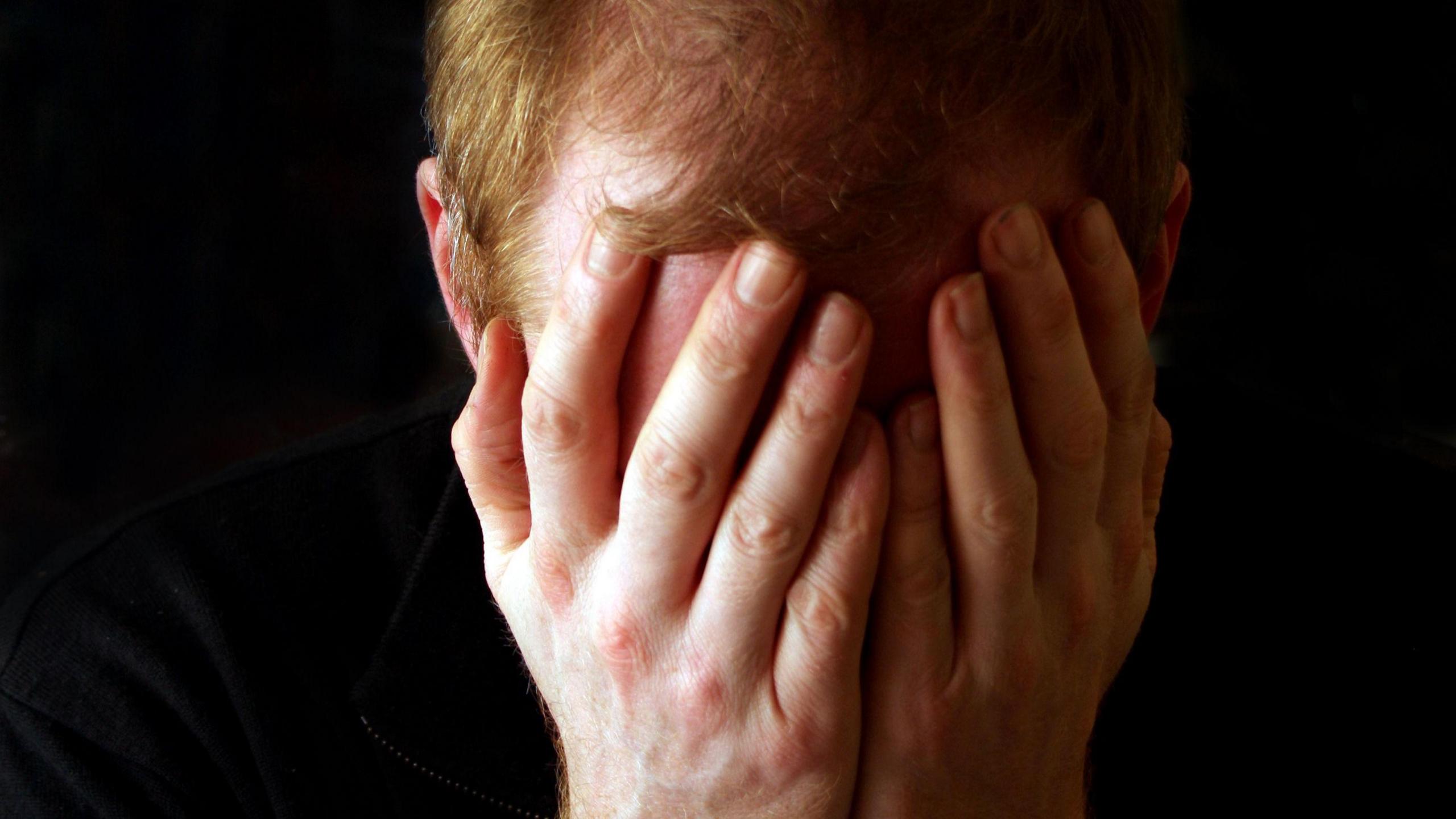 A man with thinning ginger hair holding his head in his hands, covering his face, as if sad or depressed. The background is entirely black.