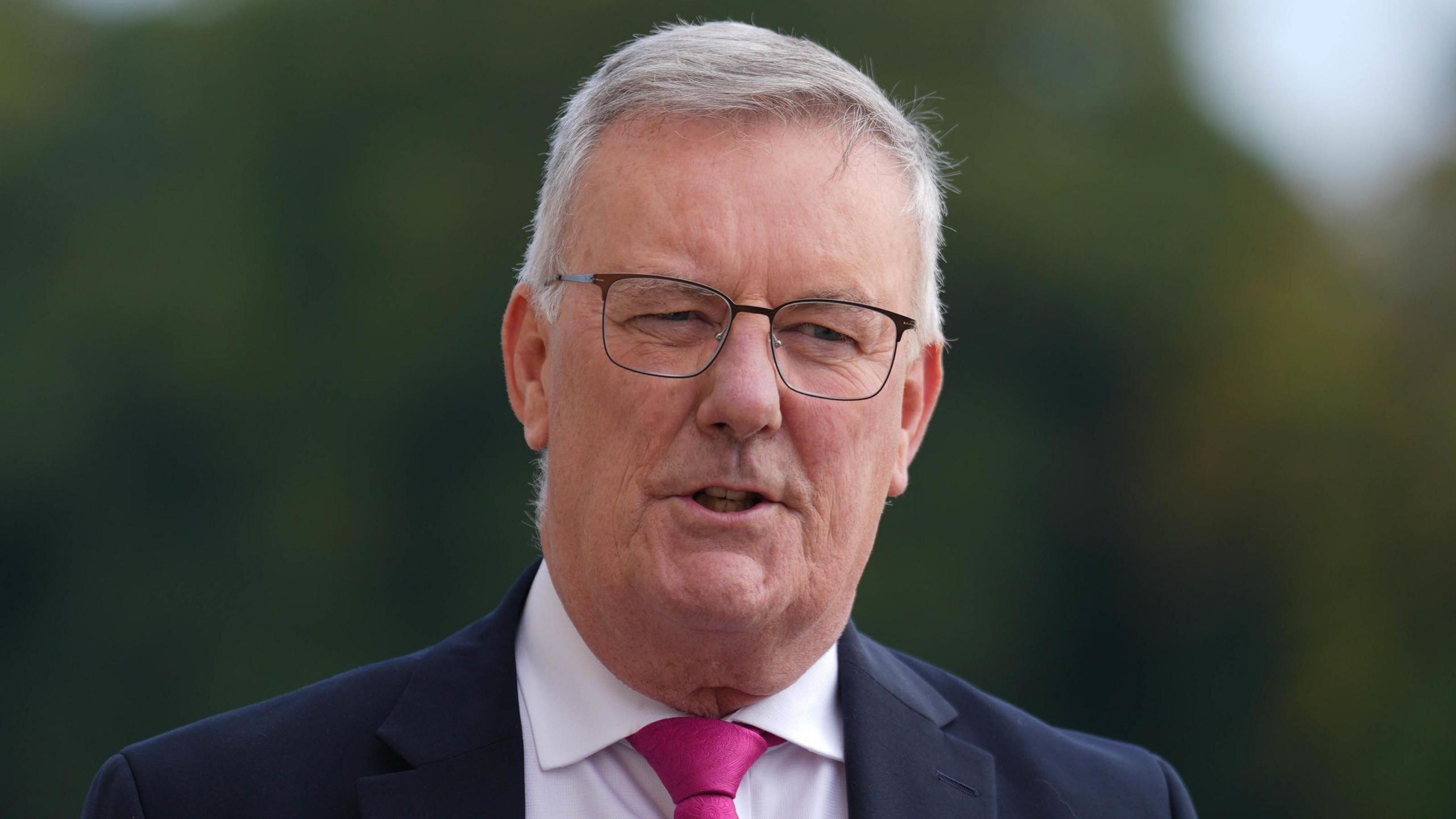 Mike Nesbitt - a man with grey hair and square-frame glasses is wearing a dark suit jacket, a white collared shirt and a pink tie. His background is blurred and he is mid-speech.