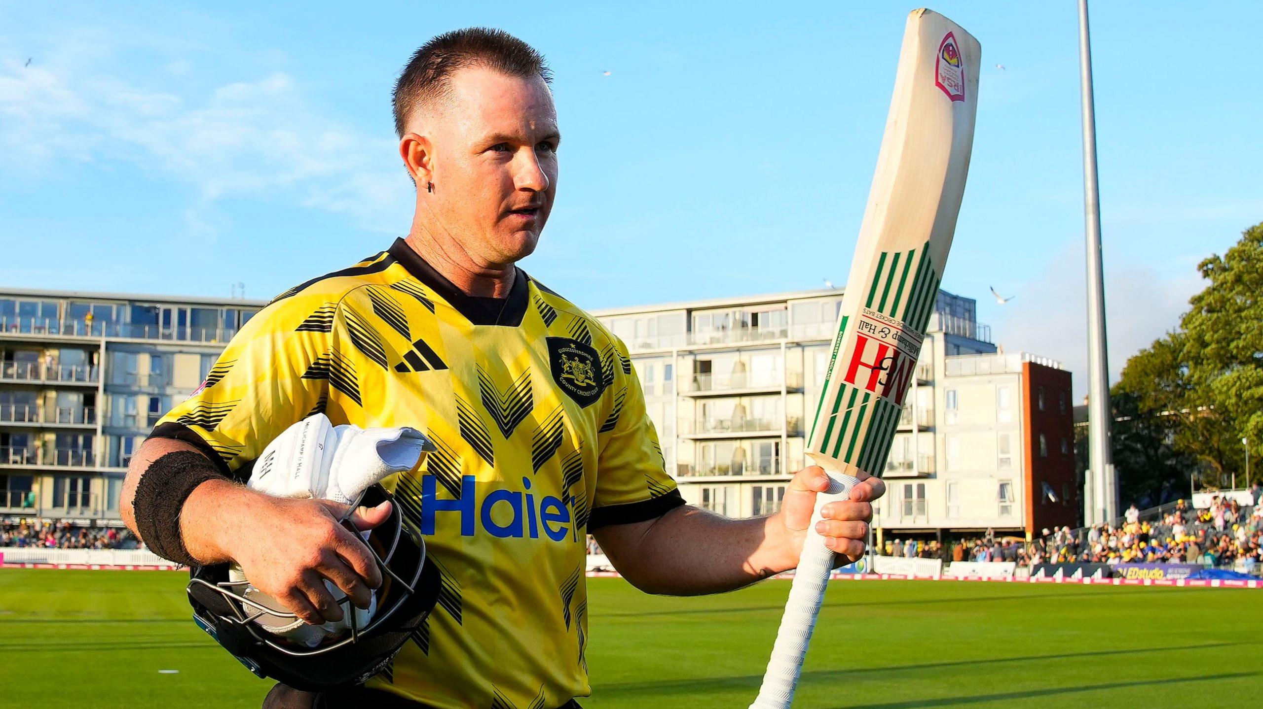 Gloucestershire's D'Arcy Short walks off the pitch at the The Seat Unique Stadium, Bristol, with his bat raised in his left hand and his helmet and gloves under his right arm.