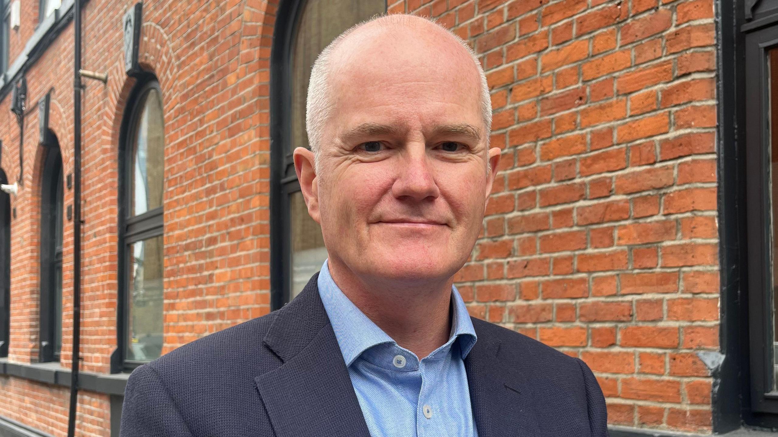 Man standing looking at the camera with a slight smile. He is wearing a shirt and suit jacket, behind him a redbrick building.