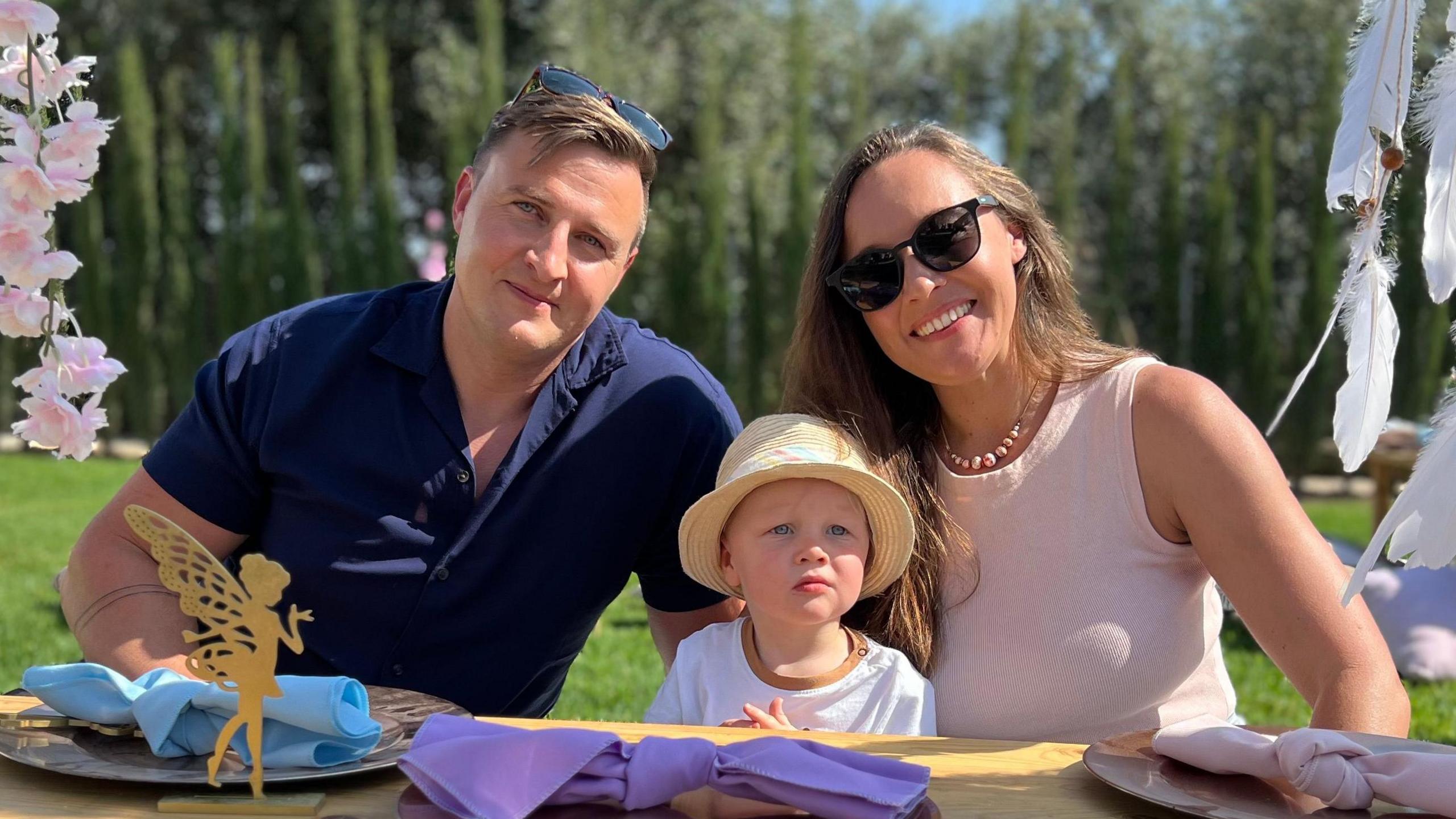 Ben Armstrong (left) sits next to his toddler son Ethan. Ben has mousy brown hair, blue eyes, and smiles at the camera. Lucy (right) sits to the right of their son and has long brown hair and smiles at the camera. Ben wears a navy shirt, with sunglasses on his head, and Ethan wears a straw sunhat, with a white t-shirt. Lucy has a pink vest top on. On the table are various colourful napkins. 