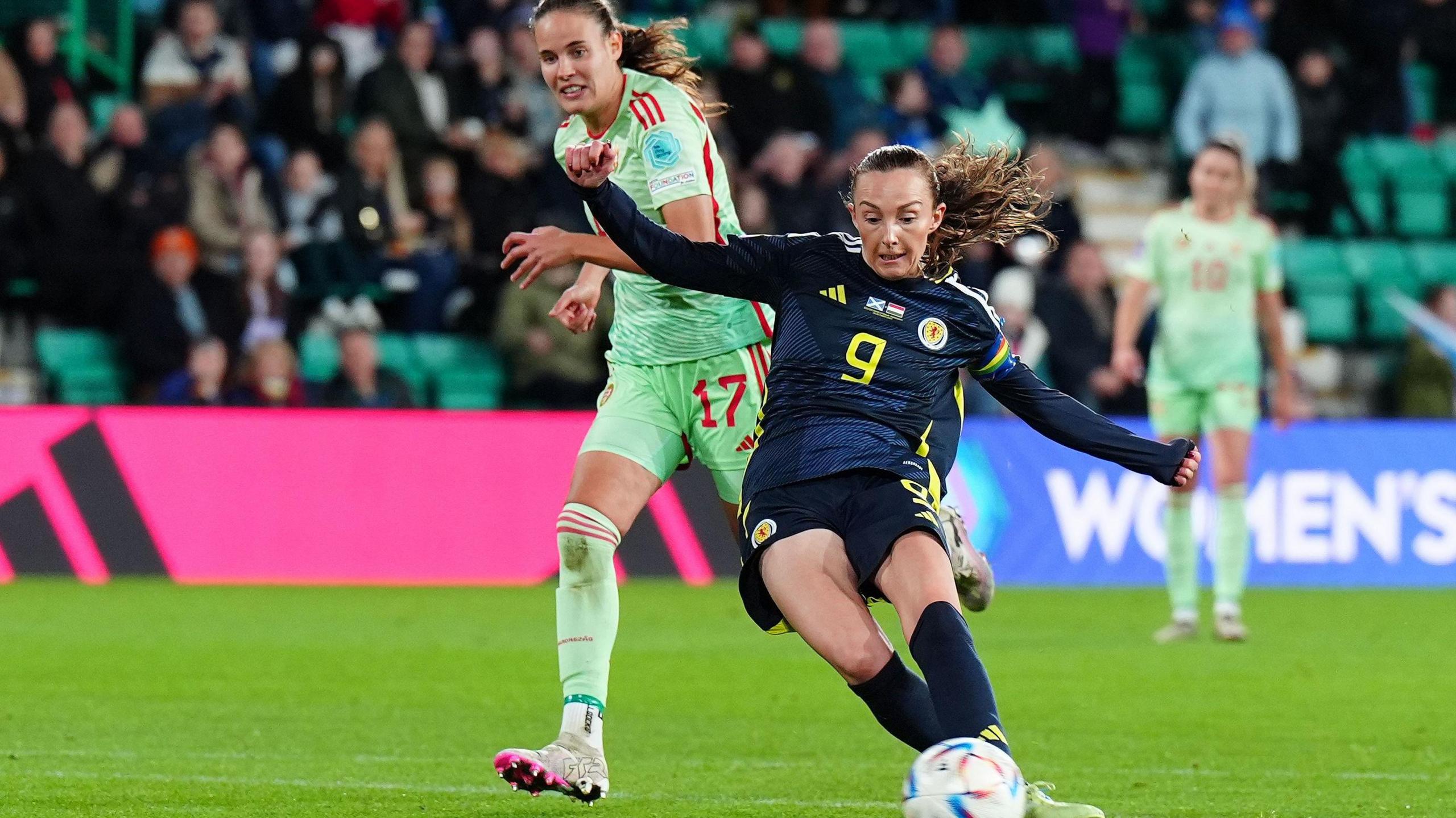 A Scotland football player kicks the ball to score whilst being chased by a Hungary player