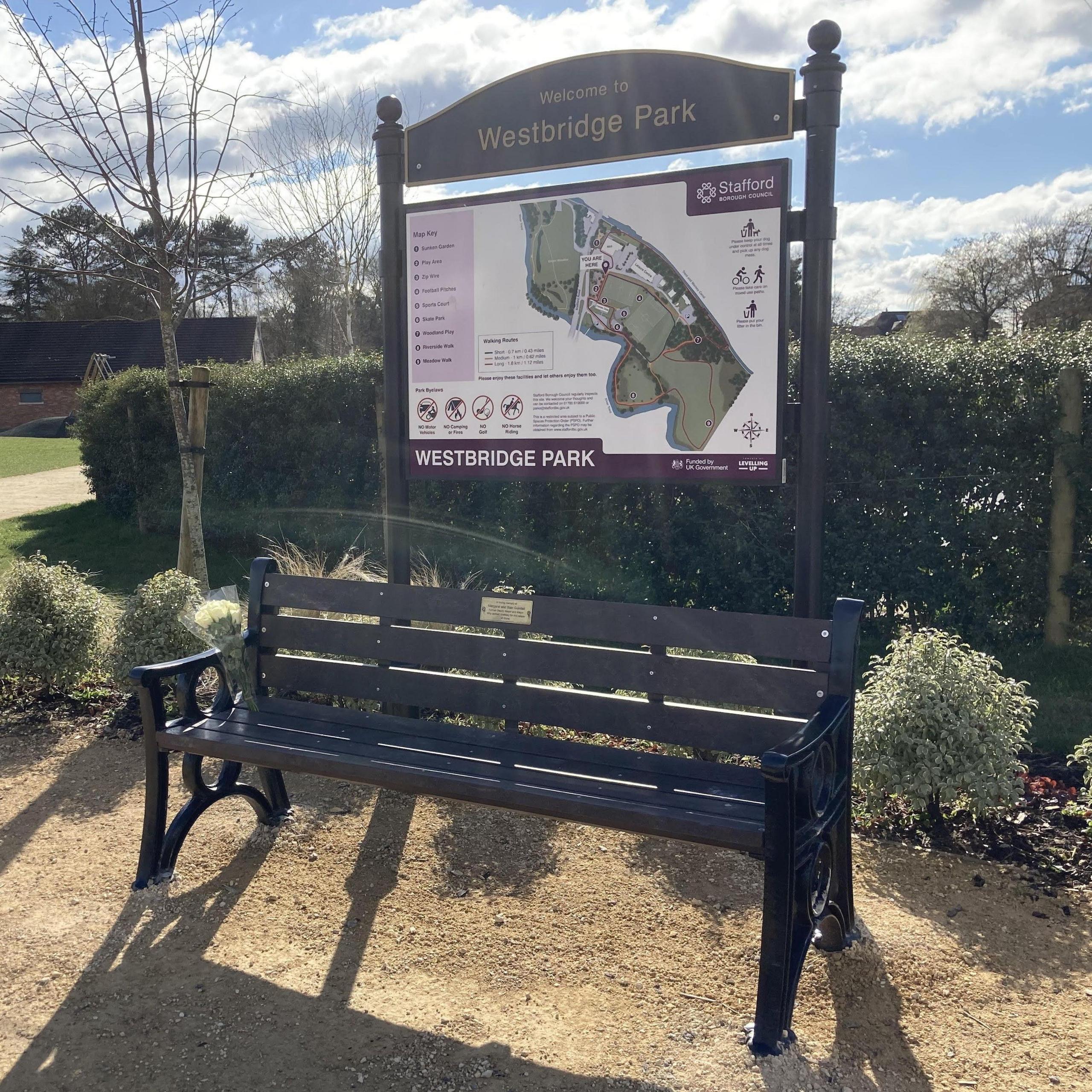 A memorial park bench in a park placed in front of a hedge and plants. A large wooden Westbridge Park sign is behind the bench featuring a map of the park.