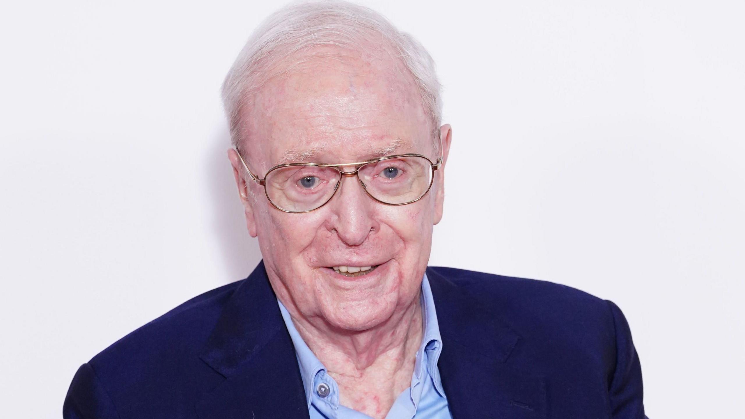 Sir Michael Caine, wearing a light blue shirt and a dark blue blazer, smiles as he is photographed at a 2023 film premiere. He is wearing gold-rimmed glasses.