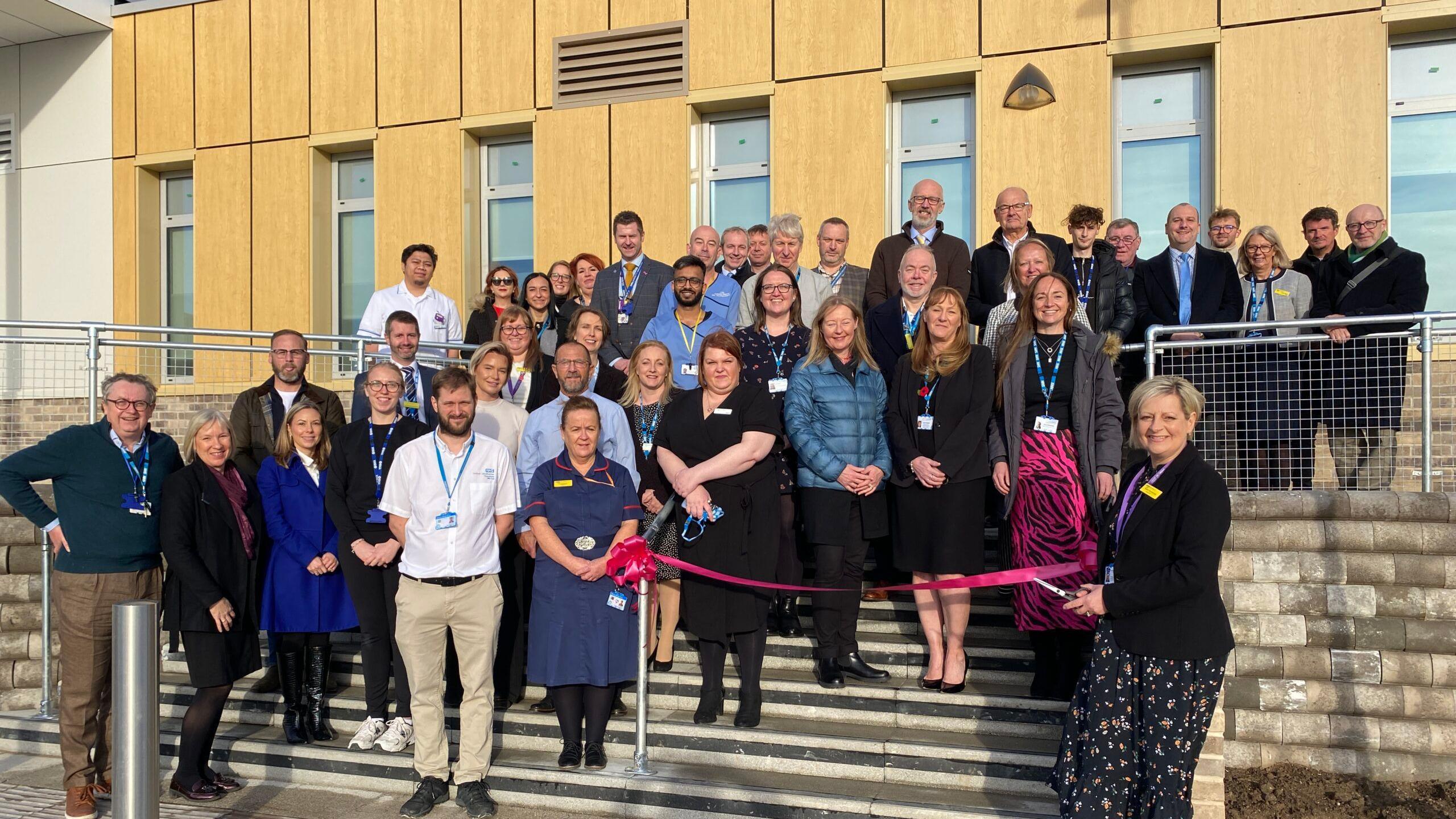 More than 40 men and women, some wearing nursing uniforms, others in smart-casual clothes, stand on the stone steps of a new health centre, which has wood-clad walls and tall windows, as a woman prepares to cut a pink ribbon. 