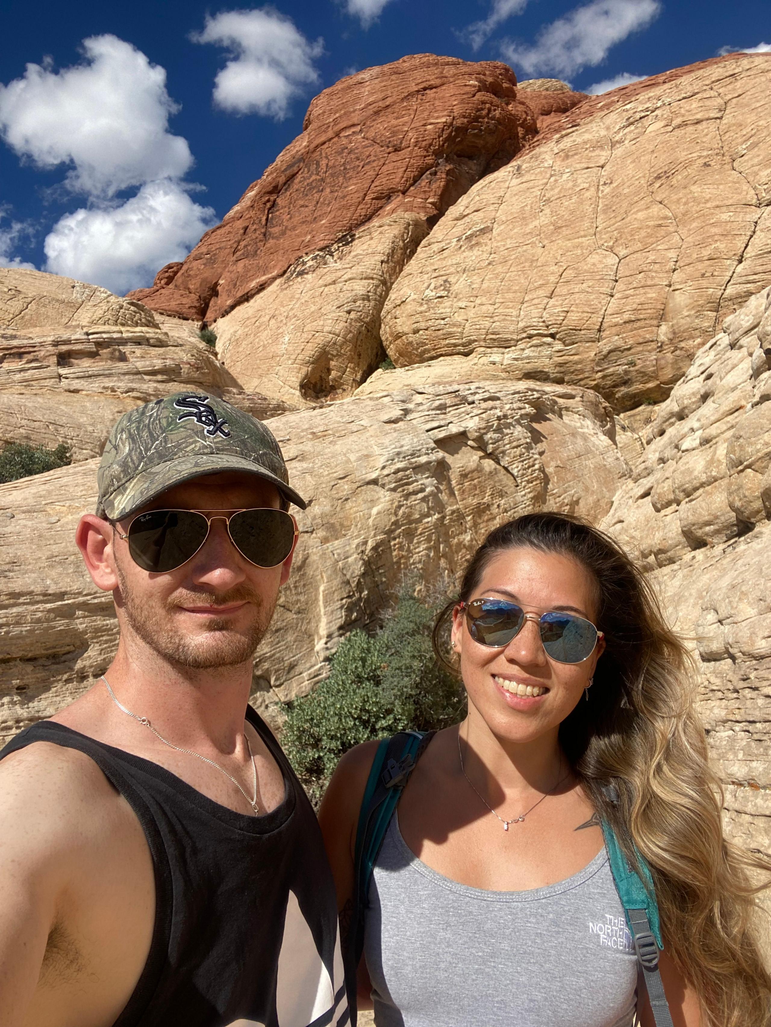 A selfie of a couple - a man and a woman - taken in front of large rocks outdoors. The woman is wearing a grey top and is smiling at the camera. The man is wearing a black tank top and a cap. Both are wearing sunglasses