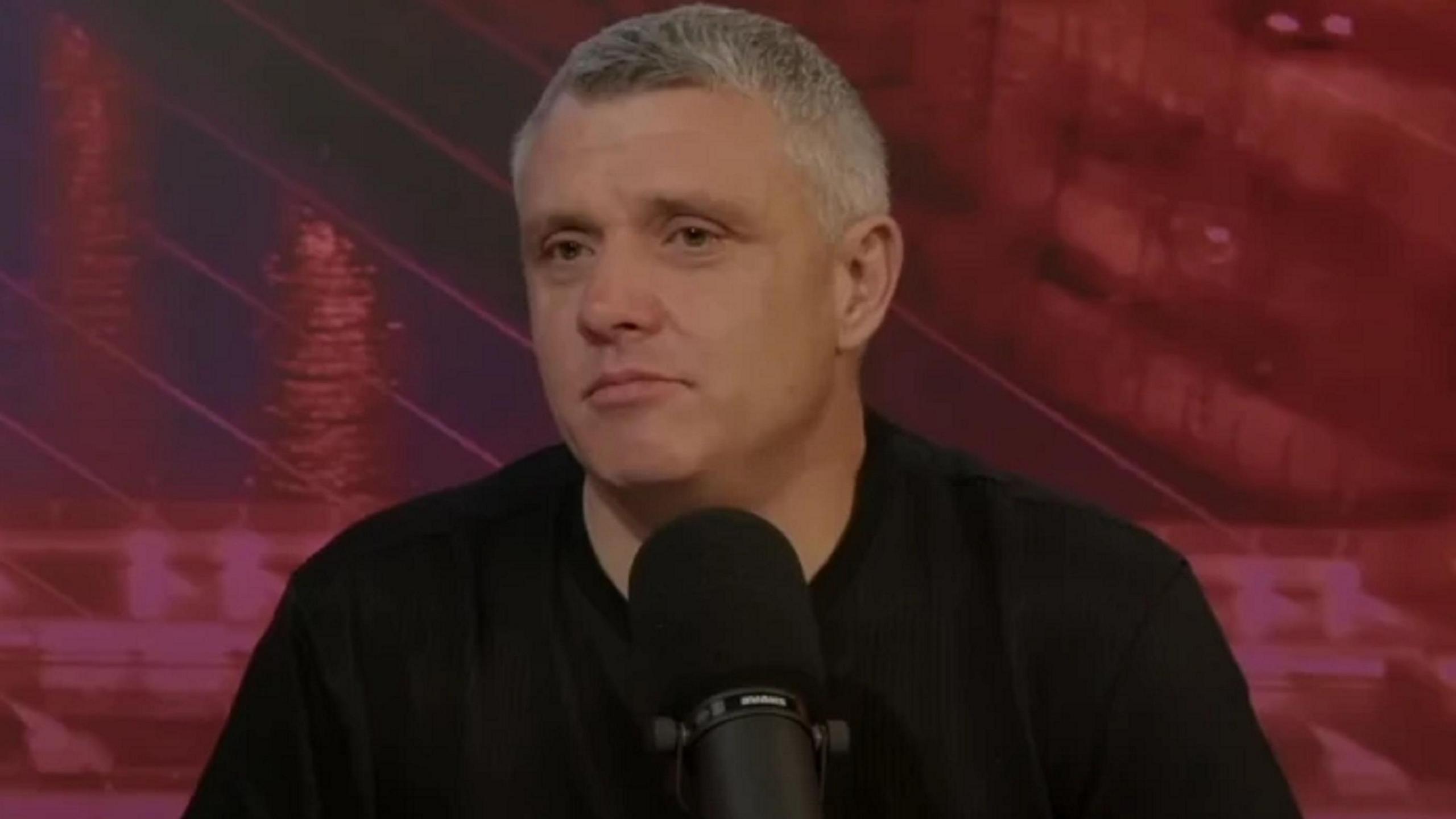 Niall Dondald has short grey hair. He is wearing a black shirt and sitting behind a microphone.